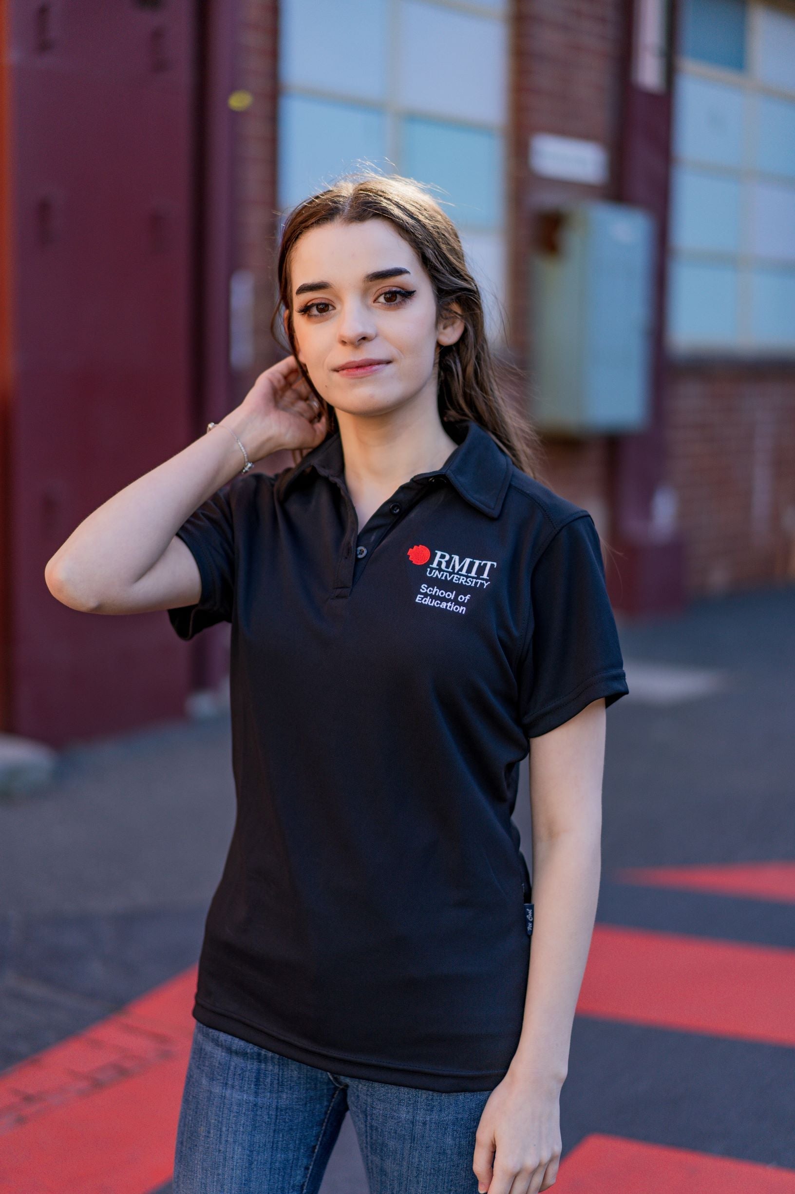 Model wearing the RMIT School of Education Polo in black, standing outdoors on the RMIT campus with buildings and paved pathways visible in the background. The model poses slightly turned to her right with her left arm relaxed by her side and her right hand gently touching her head, showing a side-front angle of the short-sleeved polo featuring “School of Education” embroidered in white on the right chest and the RMIT University logo embroidered in red and white on the left chest. Natural daylight highlight