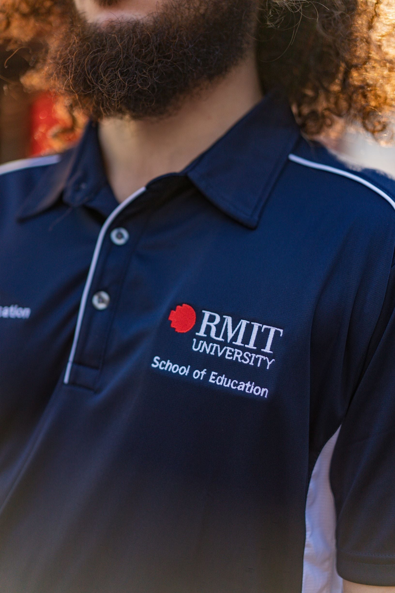 Close-up of a male model wearing the navy School of Education Polo from the RMIT Store, showing the left chest with white embroidered “School of Education” text. The polo features a collar with three buttons, the top button undone.