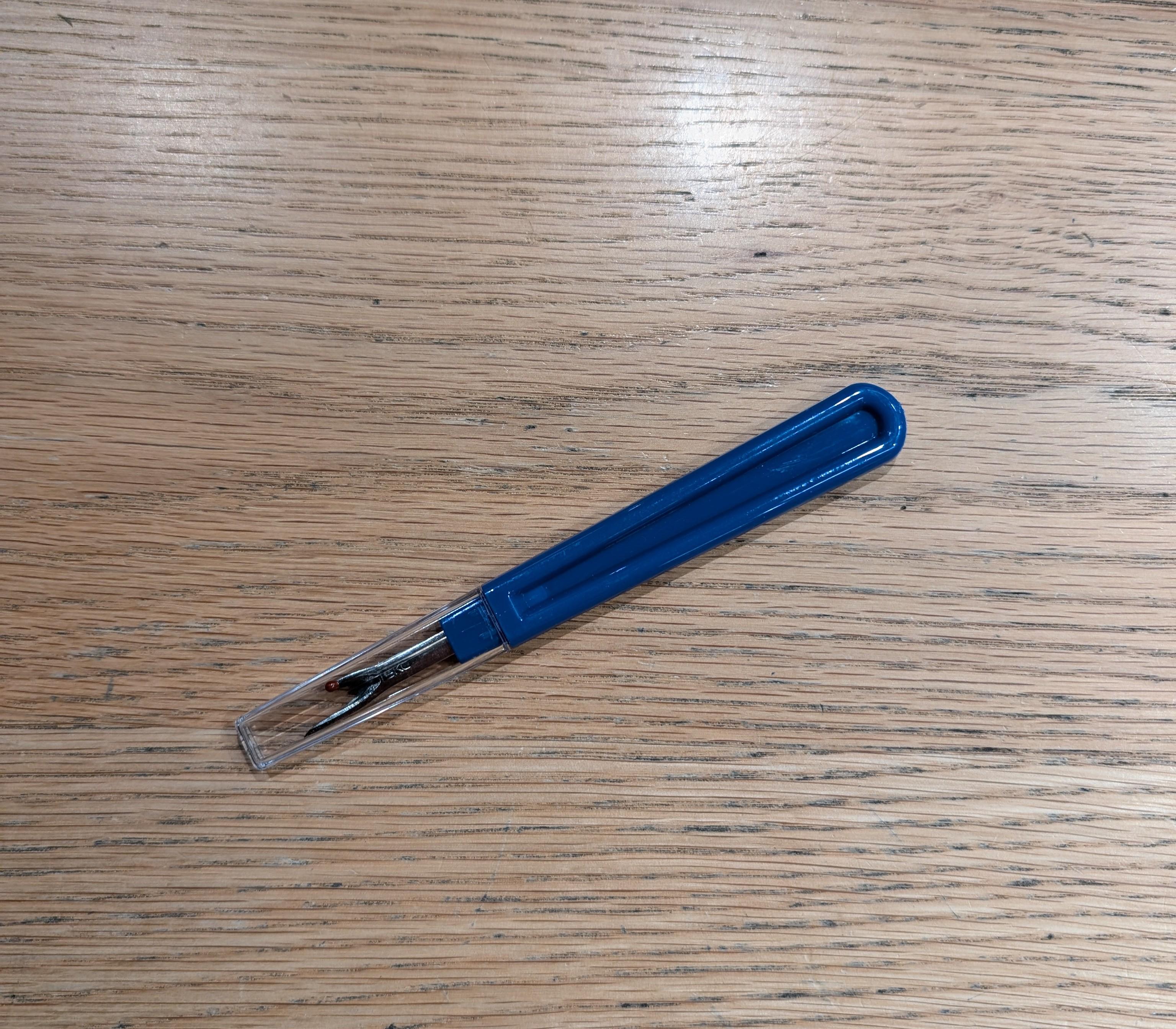 Blue seam unpicker with a clear protective cap, positioned diagonally on a wood tabletop. The plastic handle has a slightly textured grip, and the metal blade features a curved hook and pointed tip for precise thread removal. Product available at RMIT Store.