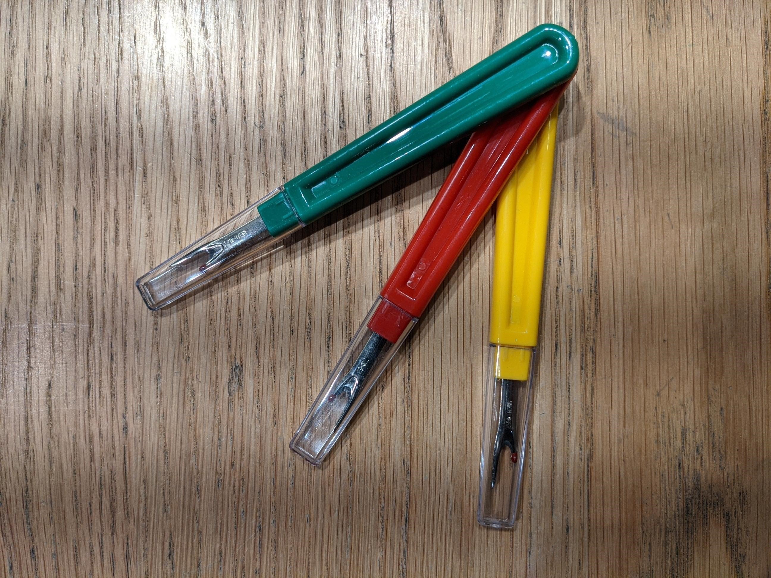 Three seam unpickers in green, red, and yellow with curved plastic handles and transparent caps, stacked one on top of the other on a wooden tabletop, each featuring a metal blade with a red safety ball tip, available from the RMIT Store.