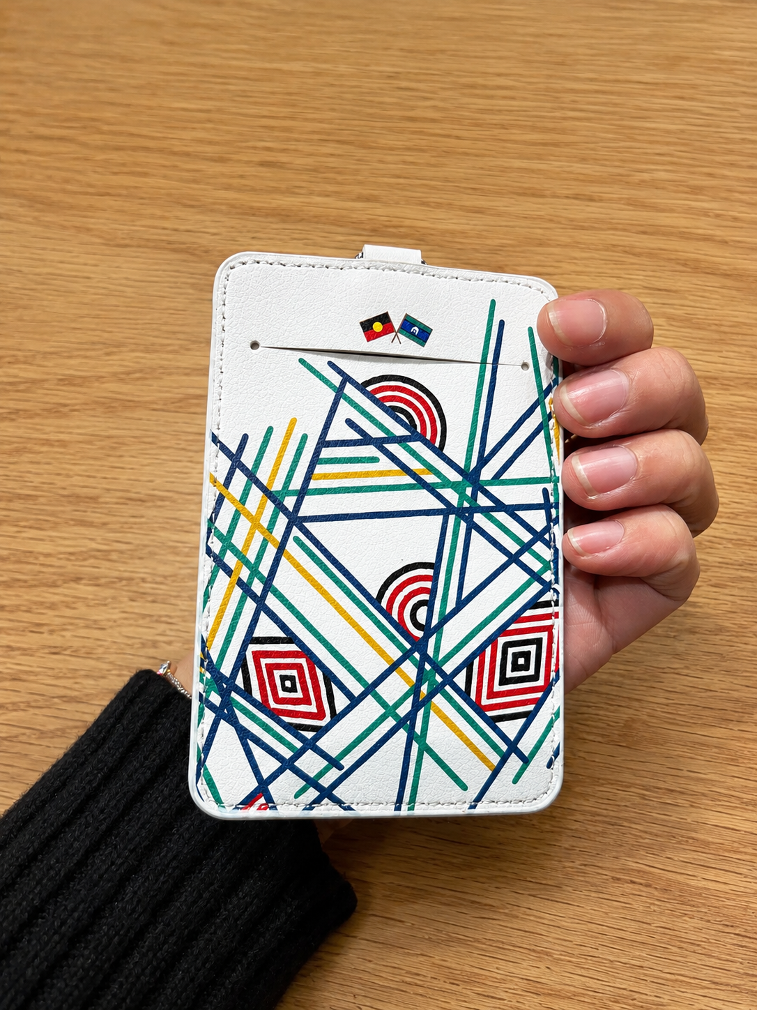 White Sentient Card Holder back view held by a model, showing colourful geometric line artwork, two small printed flags near the top card slot and white stitched edging, available at the RMIT Store.