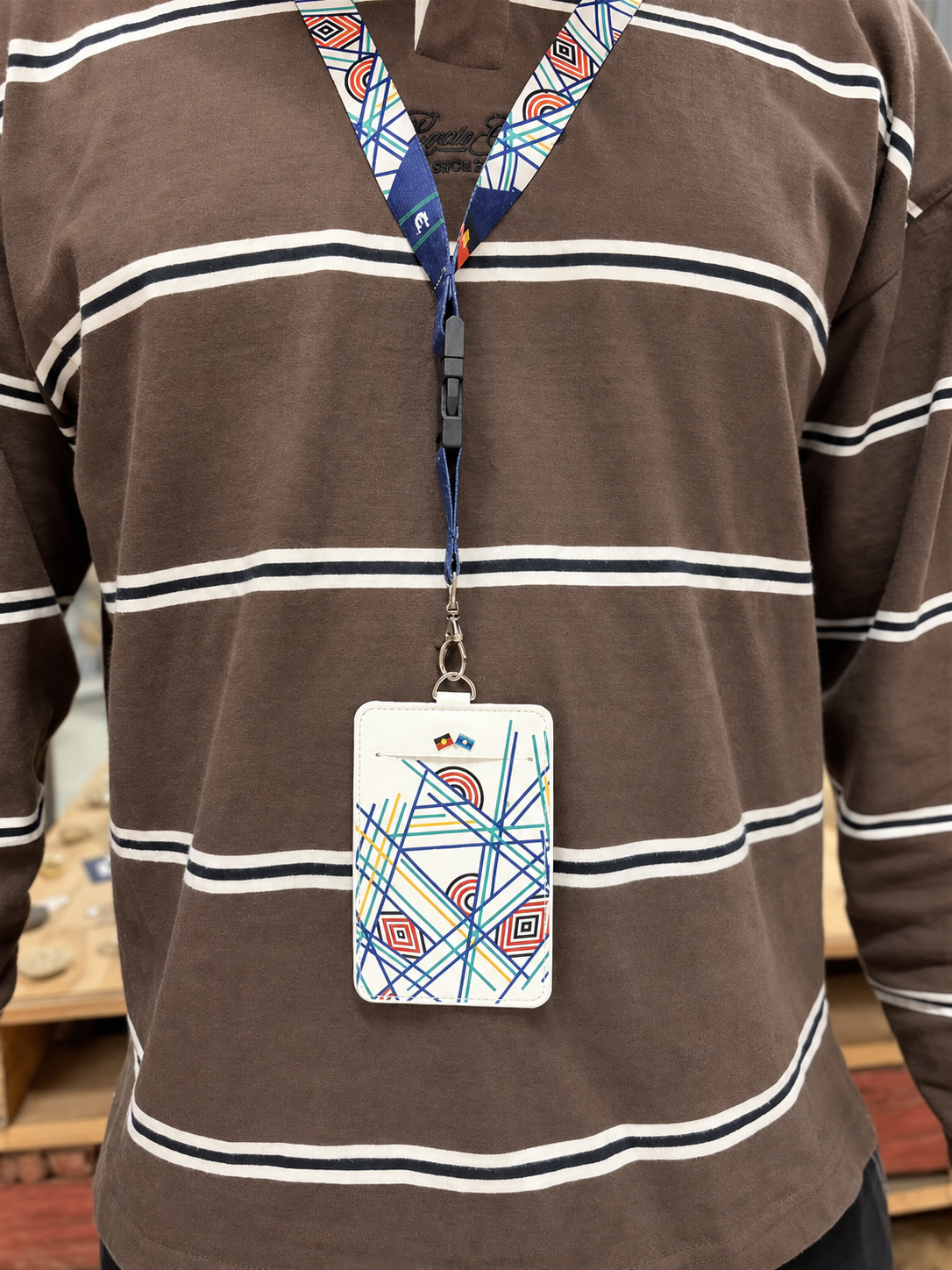 White Sentient Card Holder back view worn on a patterned RMIT lanyard by a model in a brown striped long sleeve top, showing the RMIT University logo, clear ID window and colourful geometric artwork, available at the RMIT Store.