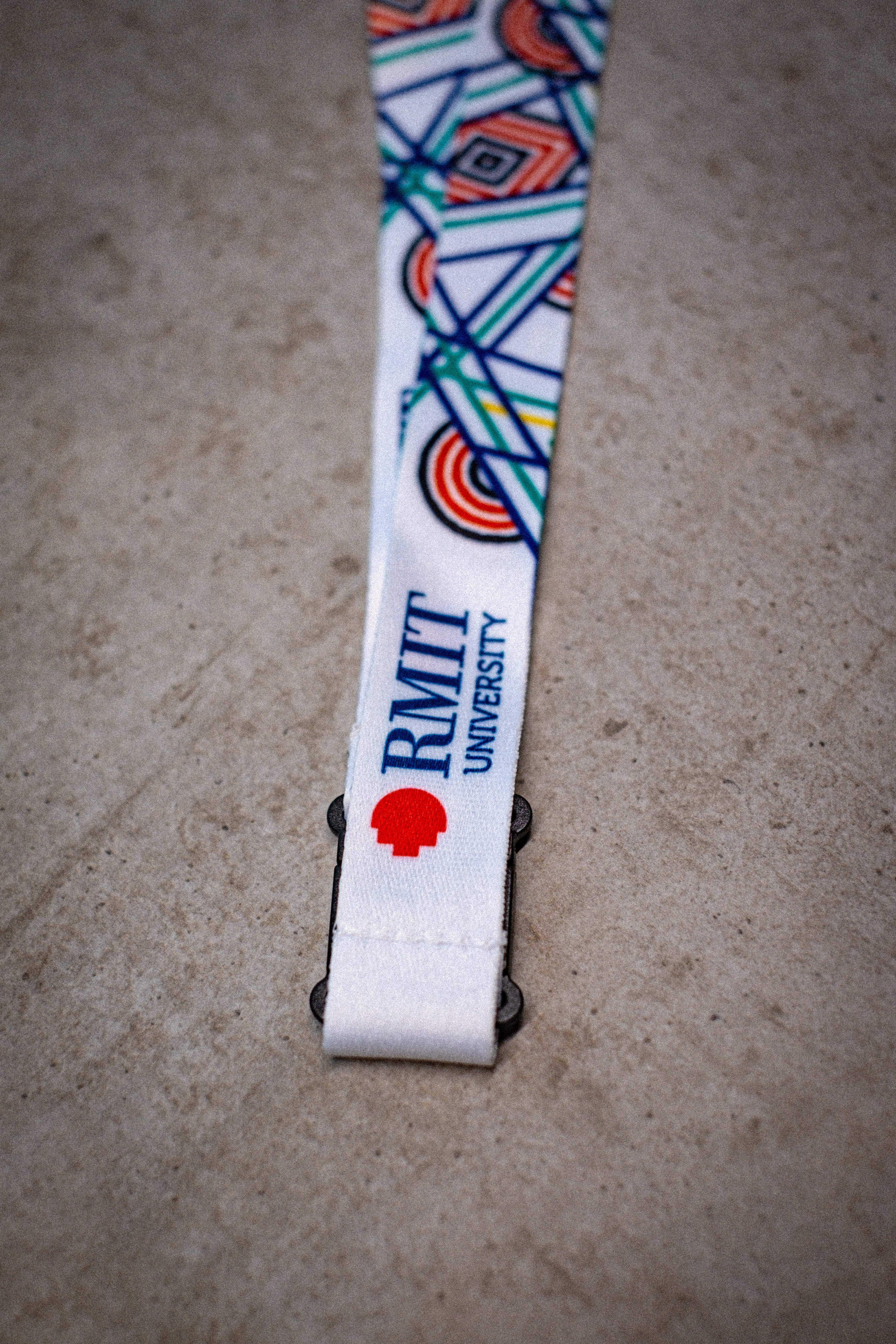 Close-up view of the Sentient Lanyard showcasing the vibrant red, yellow, green, and blue linework representing nature's evolution—sun, land, and sea. The visible section of the lanyard features the “RMIT University” print, with clear detail on the intertwining paths symbolising diverse life journeys. The lanyard is laid against a light background, highlighting the intricate artwork. Sold by RMIT Store.