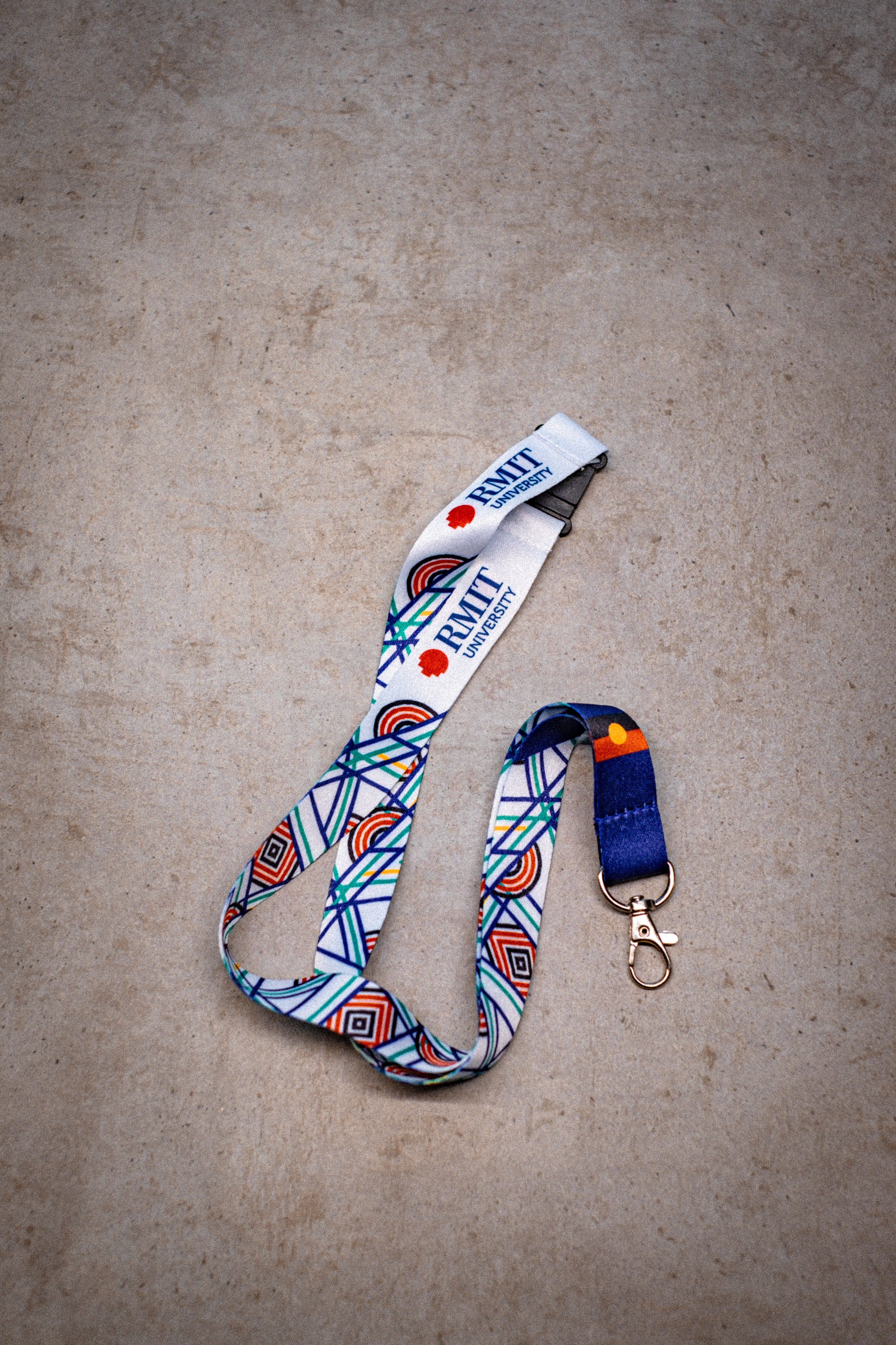 A flat-laid Sentient Lanyard featuring vibrant linework in red, yellow, green, and blue symbolising the evolution of nature—sun, land, and sea. The design includes intertwining and diverging paths, reflecting diverse life journeys. The lanyard is printed with “RMIT University” branding and shows Aboriginal and Torres Strait Islander flags. It is set on a light background for full visibility of the detailed artwork. Available at RMIT Store.