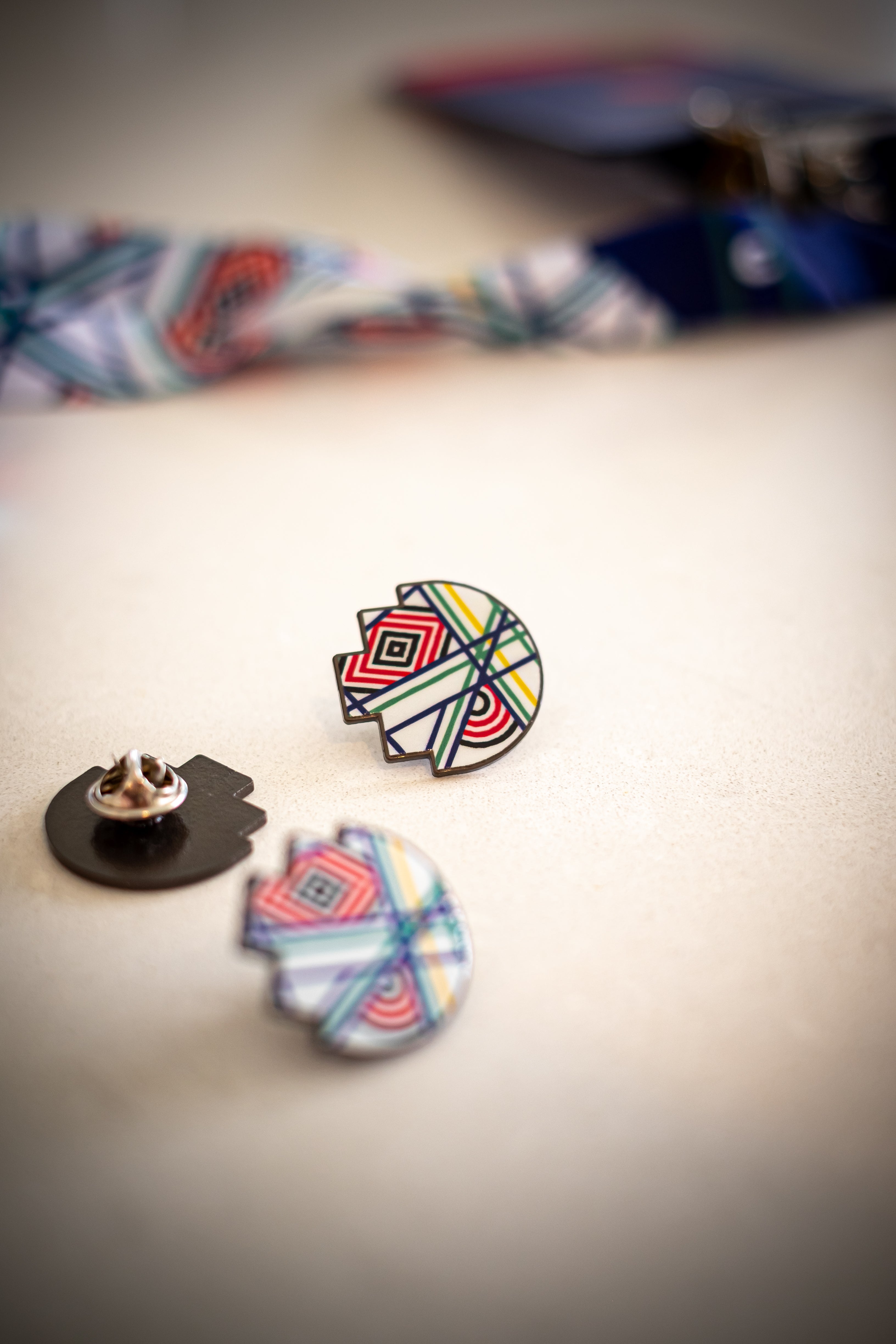 A close up view of the RMIT Store 'Sentient' lapel pin featuring vibrant geometric patterns with red, blue, yellow, and green intersecting lines. The pin is displayed on a light surface against a blurred 'Sentient' lanyard, highlighting its intricate design and metal clasp backing.