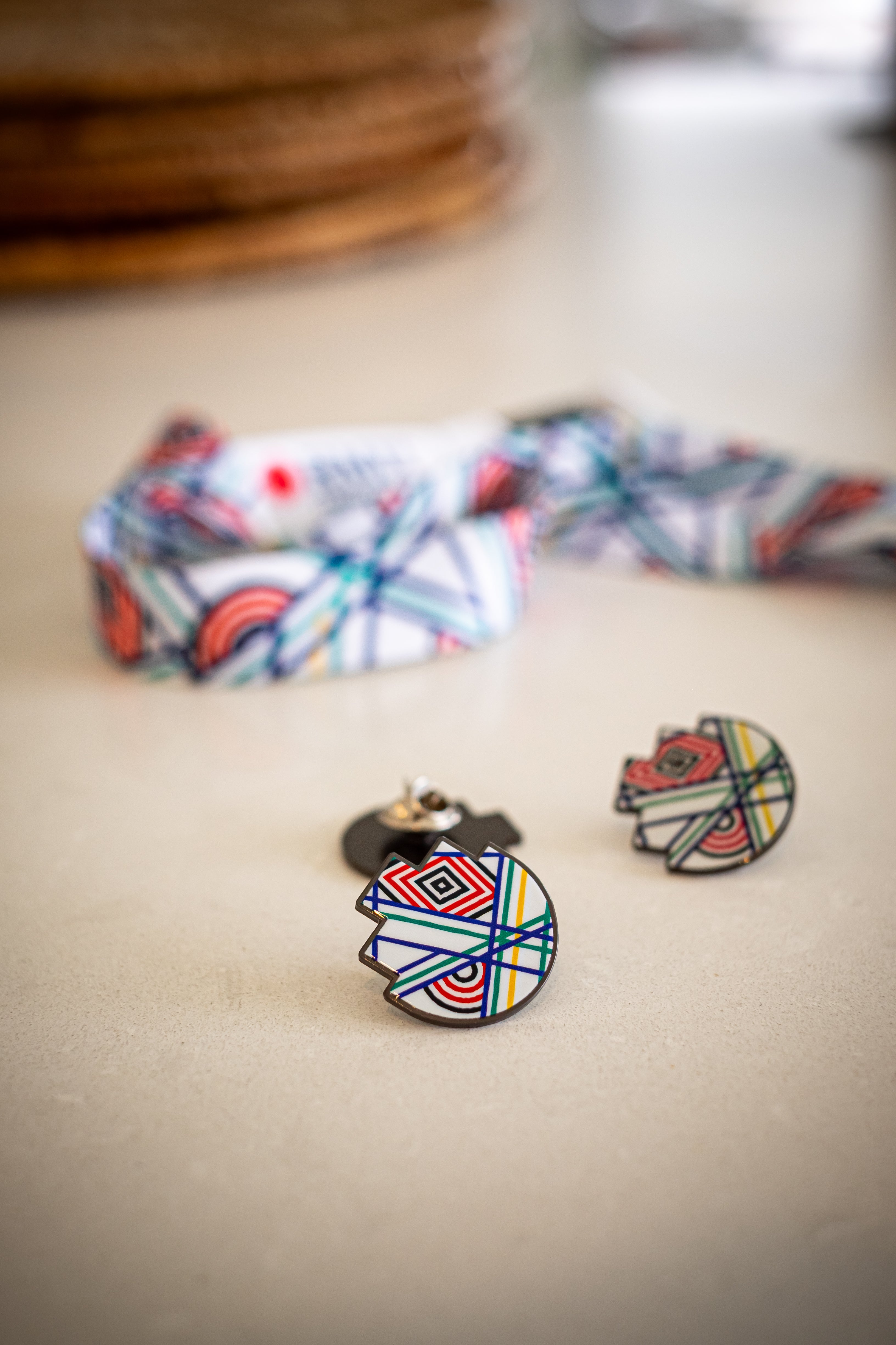 RMIT Store 'Sentient' lapel pin placed on a desk, showing colourful geometric patterns. A blurred 'Sentient' lanyard in the background complements the pin design, emphasising its artistic details and sharp metal finish.