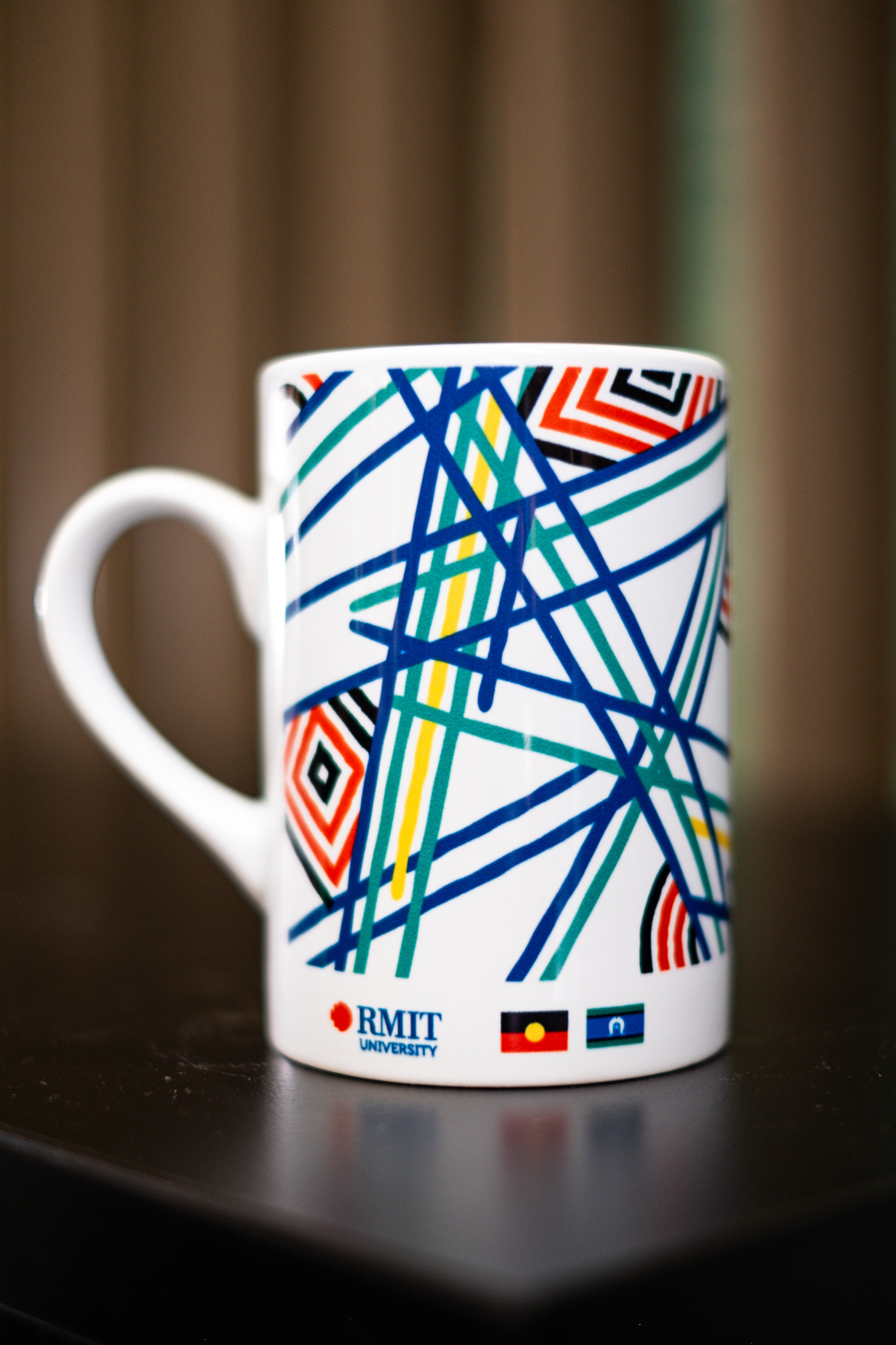 RMIT Store Sentient Mug, white ceramic, standing upright on a black tabletop with the handle positioned to the left. The entire Sentient artwork wraps around the body of the mug in vibrant red, blue, yellow, and green linework symbolising the sun, land, and sea. The design illustrates life’s varied and interconnected journeys through continuous and broken lines. At the bottom of the mug, the RMIT University logo is printed alongside the Aboriginal and Torres Strait Islander flags, reinforcing the mug’s cult