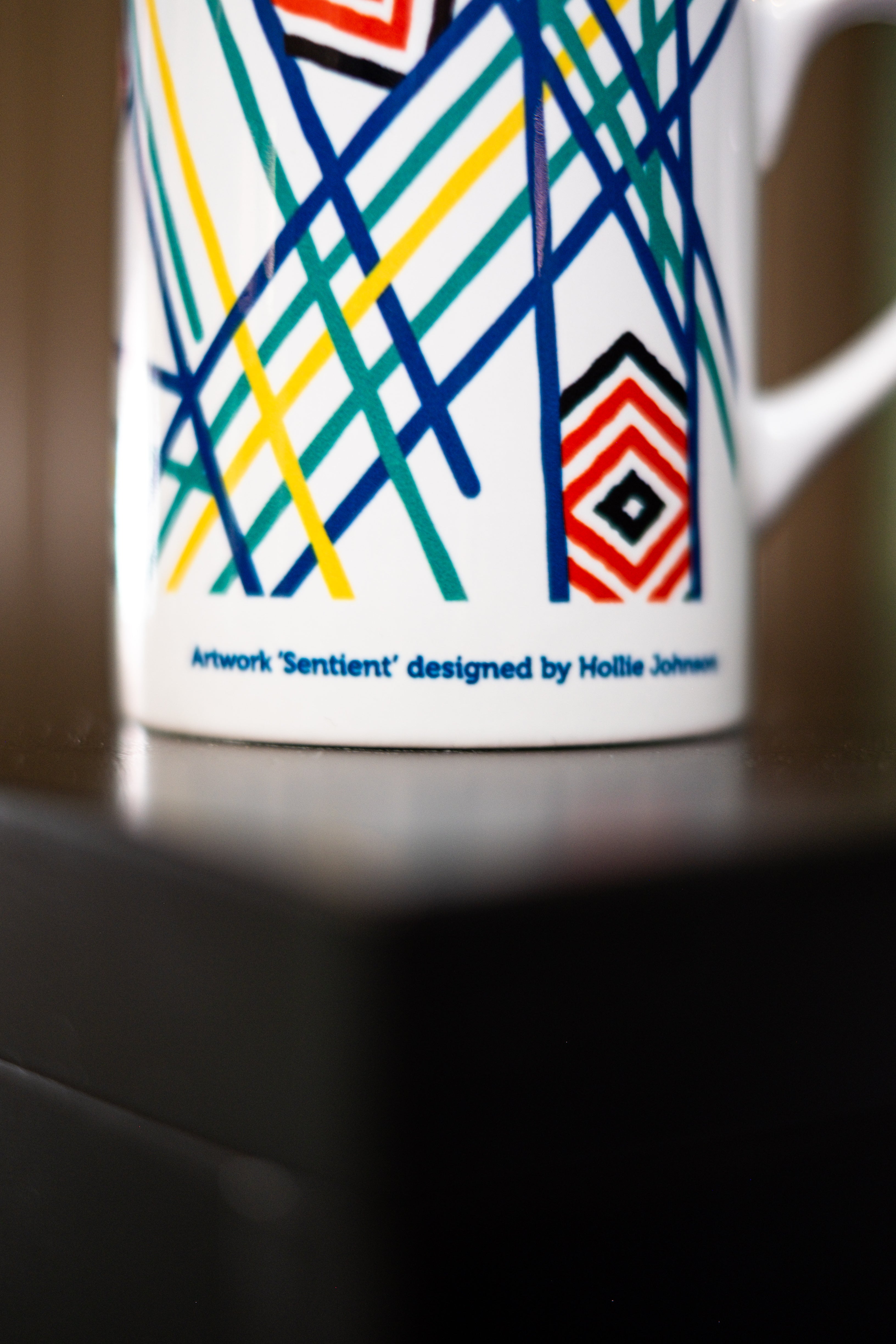 Close-up view of the base of the RMIT Store Sentient Mug, white ceramic, showing the printed text in small black font that reads: Artwork 'Sentient' designed by Hollie Johnson. The mug is placed on a black tabletop,