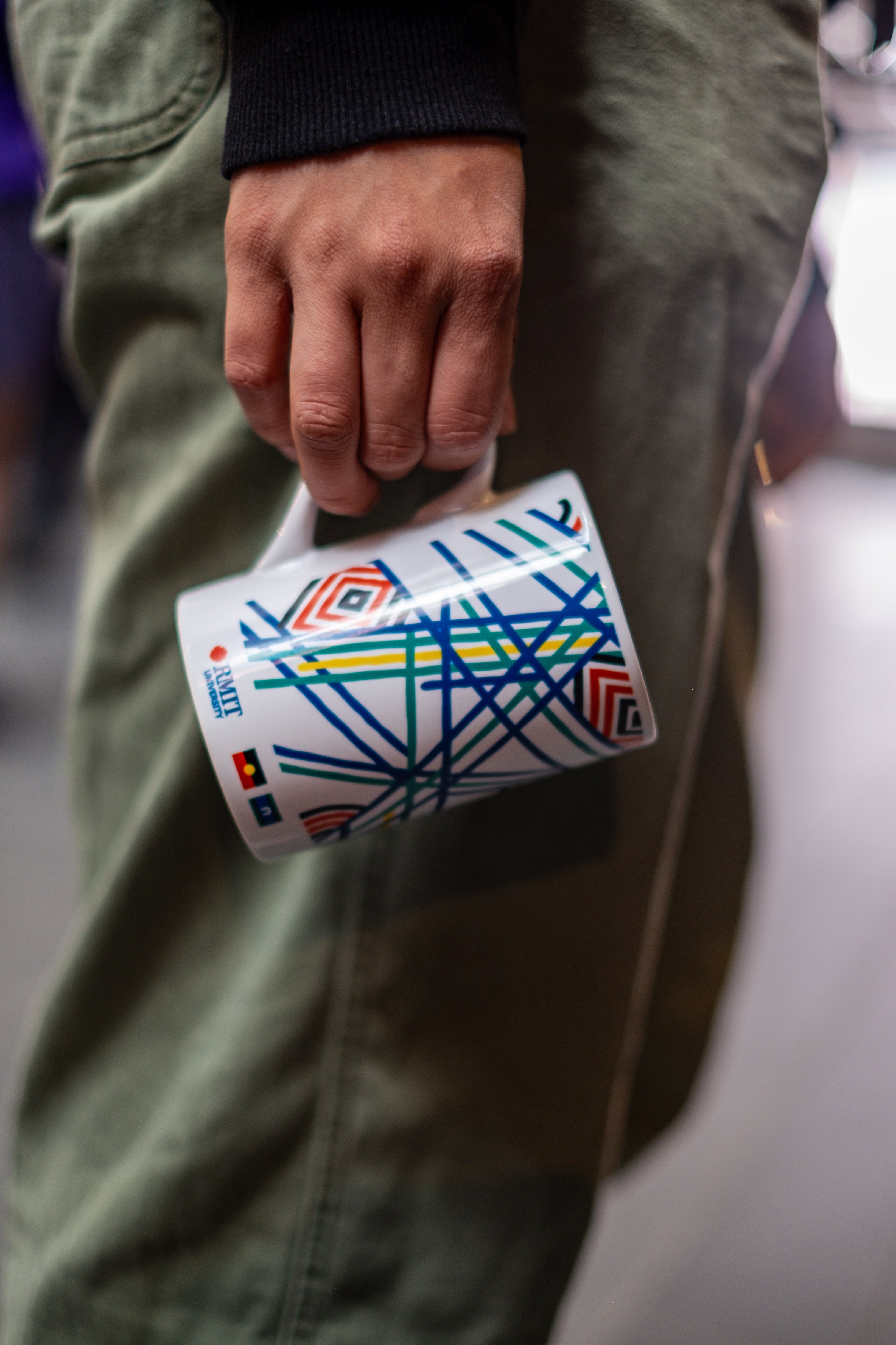 RMIT Store Sentient Mug, held by a model with their right hand, positioned to the right of the mug’s handle. The white ceramic mug features vibrant, multicoloured Sentient artwork across its surface. The intricate linework, in shades of red, blue, yellow, and green, reflects the evolution of nature, symbolising sun (yellow), land (green), and sea (blue). The RMIT University  and Indigenous flags prints are visible on the mug, subtly located along the body. 