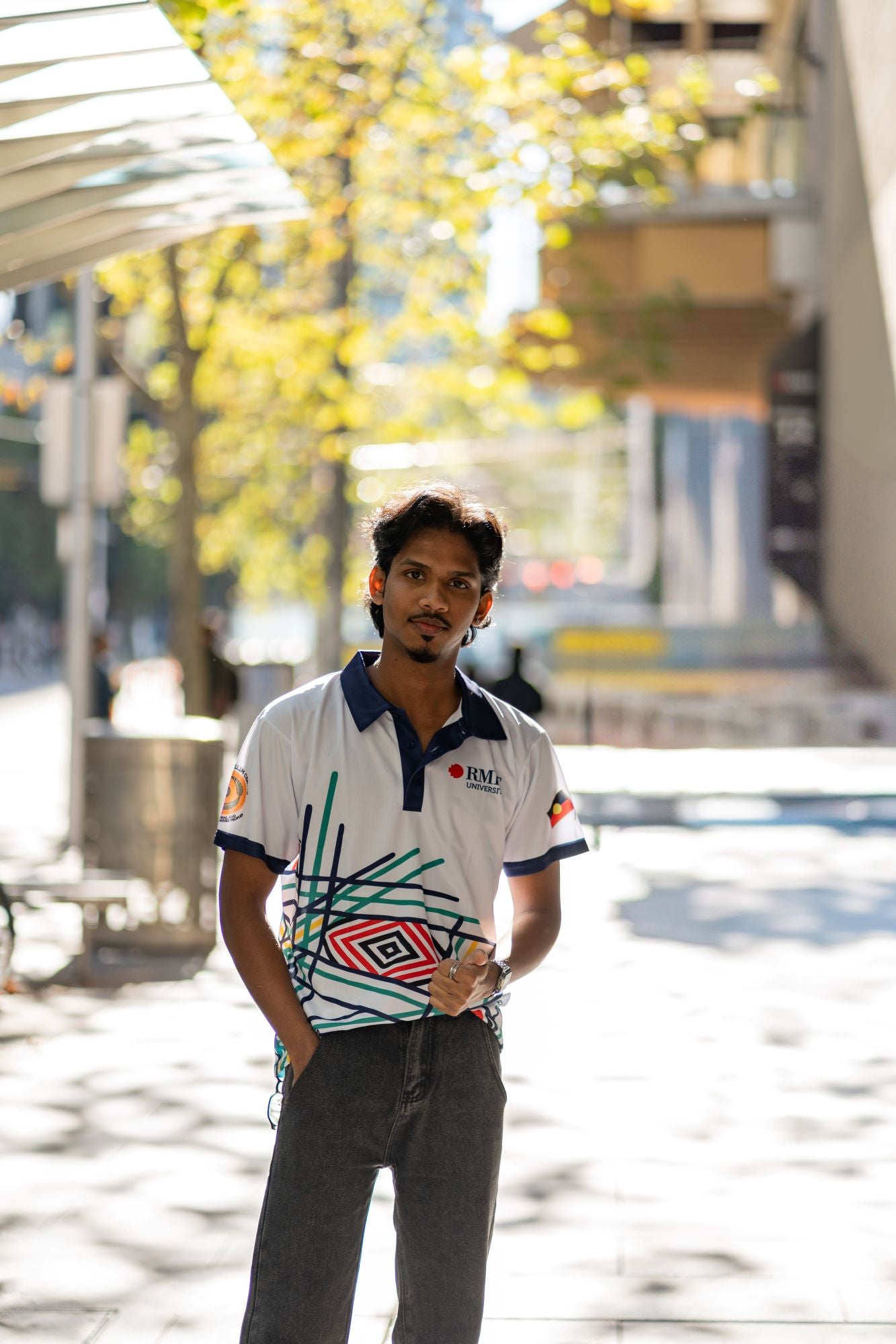Front view of male model wearing the Sentient Polo with a navy collar and Indigenous 'Sentient' artwork by Hollie Johnson, styled with denim jeans, one hand in pocket and the other giving a thumbs up, photographed in an urban setting. Available at the RMIT Store.