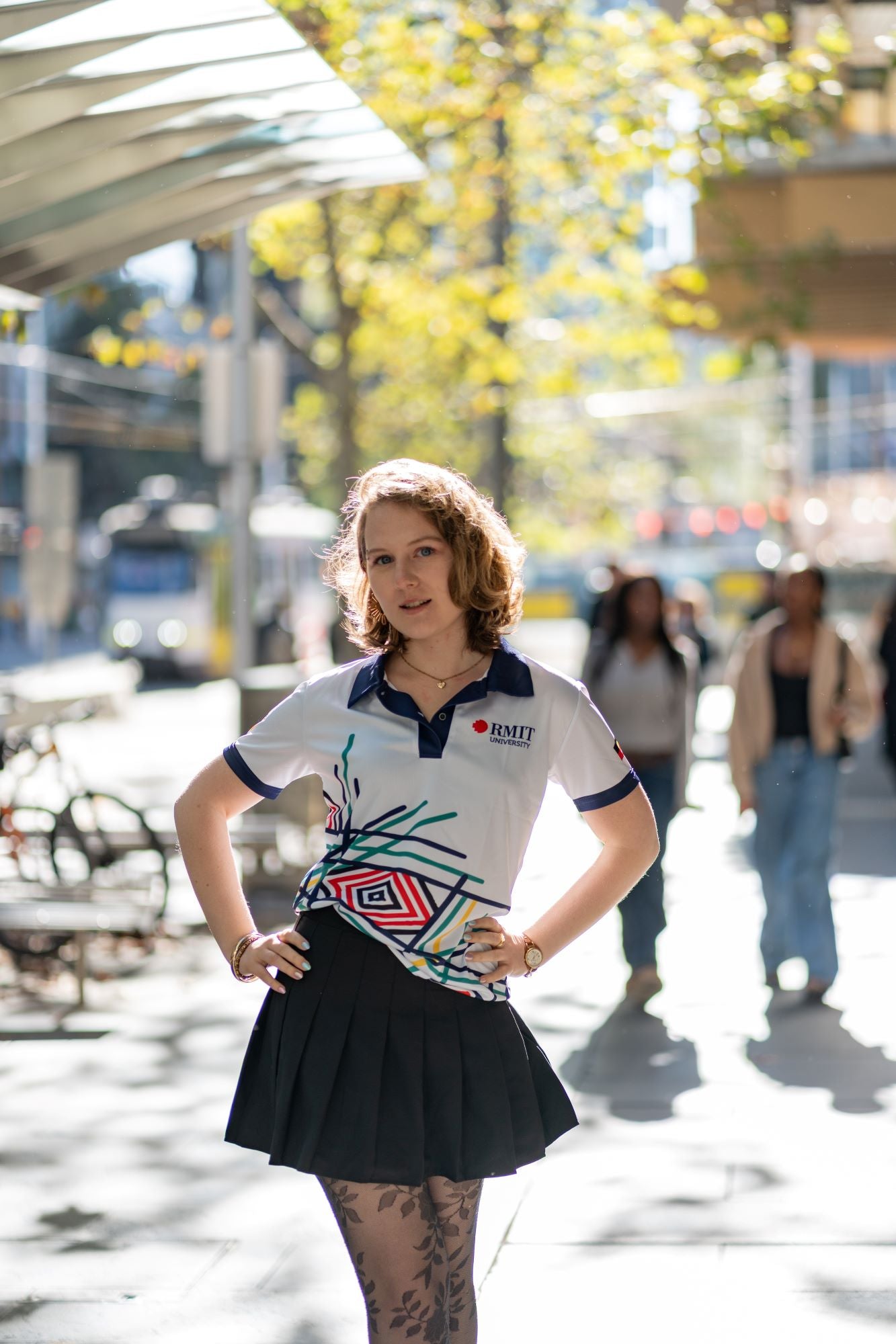 Female model wearing the Sentient Polo with navy collar and Indigenous 'Sentient' artwork by Hollie Johnson, standing in an urban setting with hands on hips and styled with a black skirt; front view highlighting the fitted cut and patterned fabric. Available at the RMIT Store.