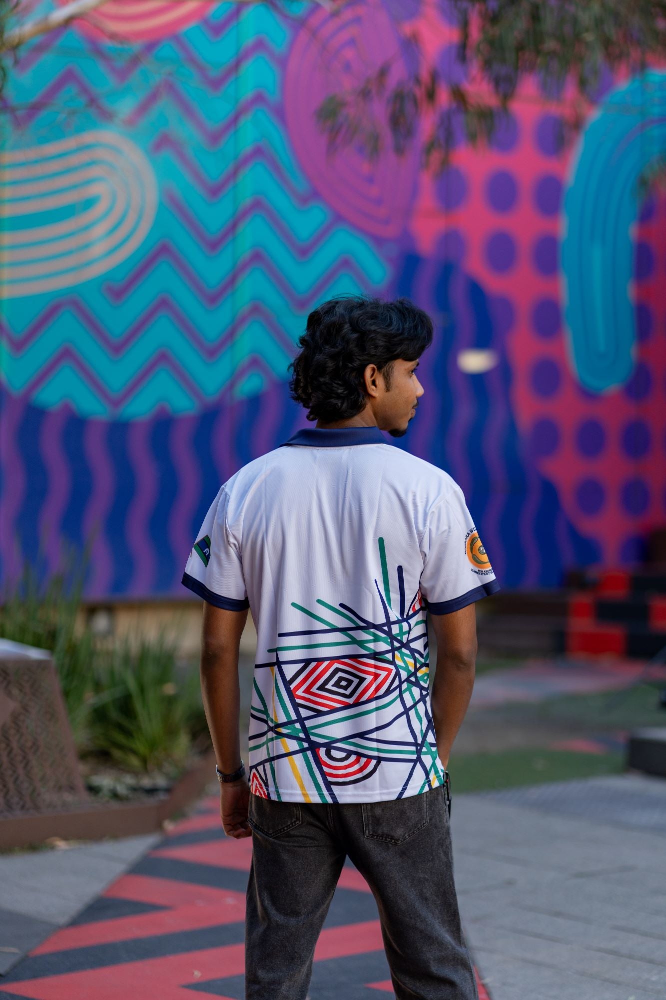 Rear view of male model wearing the Sentient Polo with a navy collar and Indigenous 'Sentient' artwork by Hollie Johnson, set against a colourful urban background. Available at the RMIT Store.