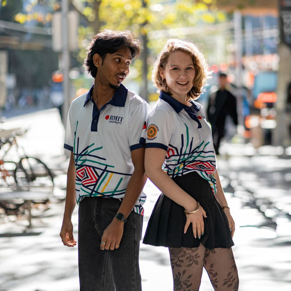 Male and female models wearing Sentient Polos featuring Hollie Johnson’s Indigenous 'Sentient' artwork with navy collars; models stand back-to-back in an urban setting, the female model in a black skirt looking into the camera, and the male model in jeans looking at her. Available at the RMIT Store.