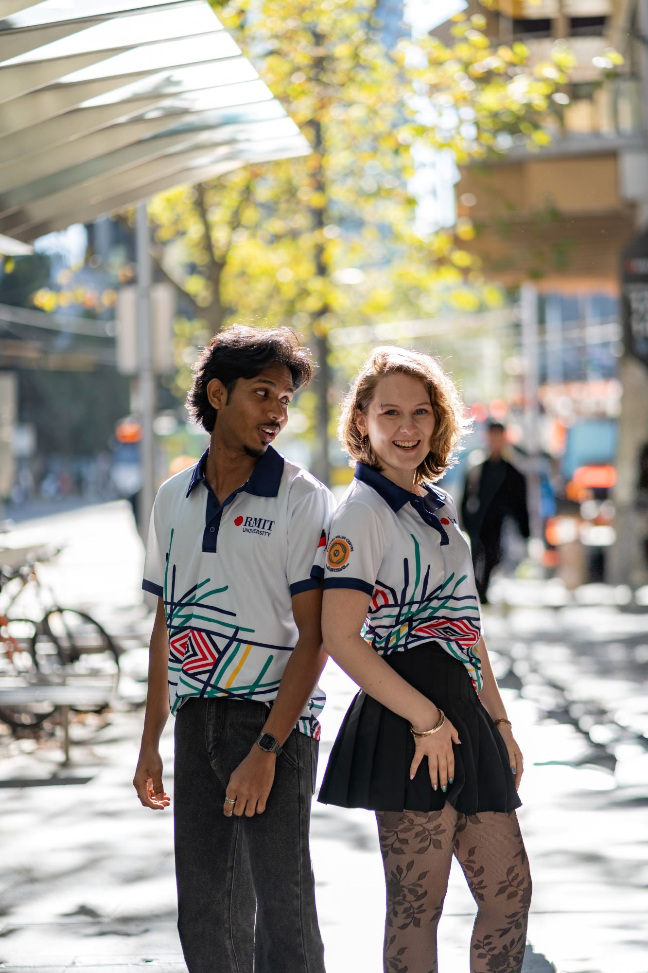 Male and female models wearing Sentient Polos featuring Hollie Johnson’s Indigenous 'Sentient' artwork with navy collars; models stand back-to-back in an urban setting, the female model in a black skirt looking into the camera, and the male model in jeans looking at her. Available at the RMIT Store.