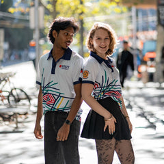 Male and female models wearing Sentient Polos featuring Hollie Johnson’s Indigenous 'Sentient' artwork with navy collars; models stand back-to-back in an urban setting, the female model in a black skirt looking into the camera, and the male model in jeans looking at her. Available at the RMIT Store.