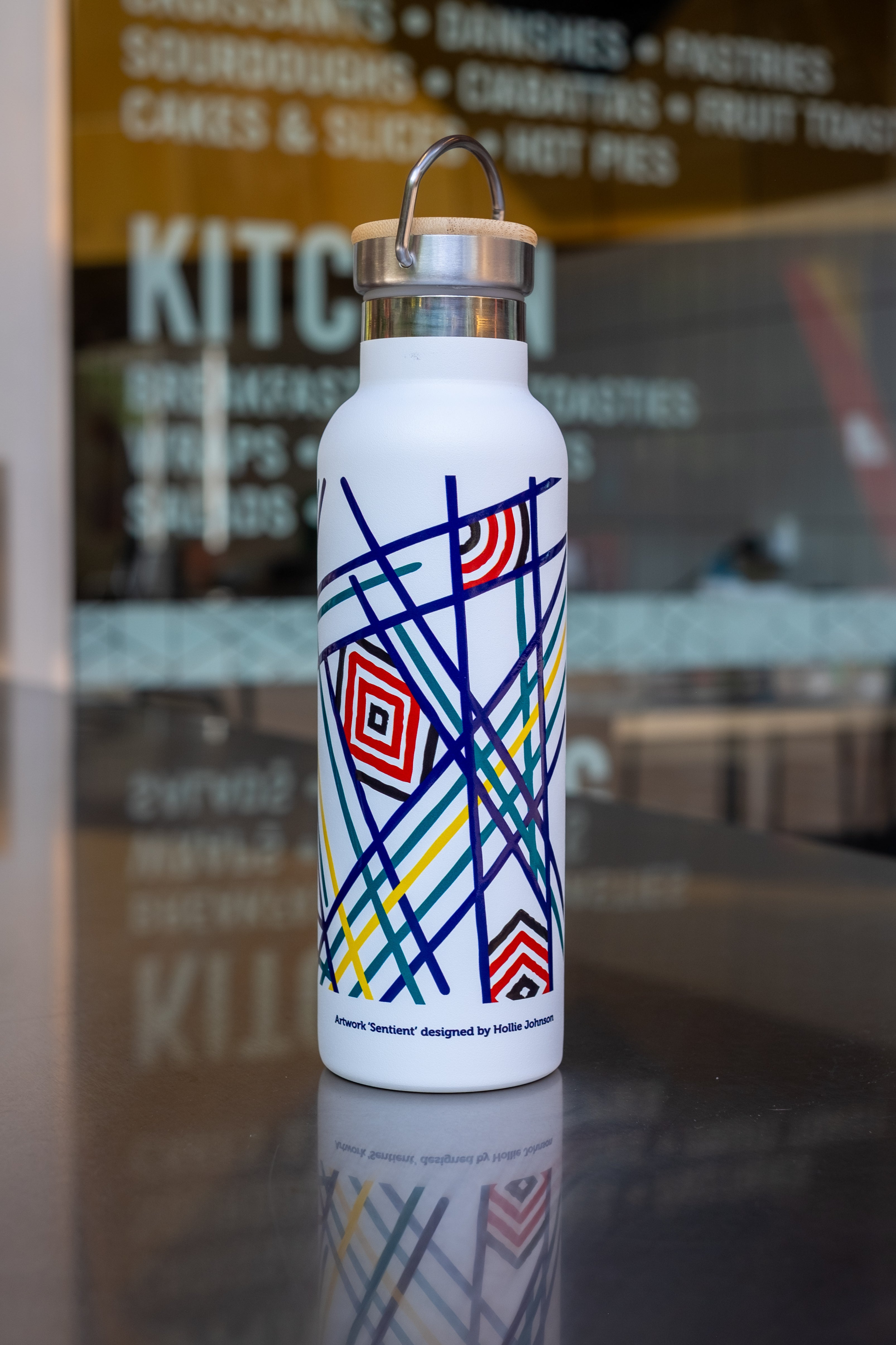 White thermo bottle showcasing geometric artwork titled 'Sentient' by Hollie Johnson, positioned against a bakery and kitchen backdrop, on a reflective surface.