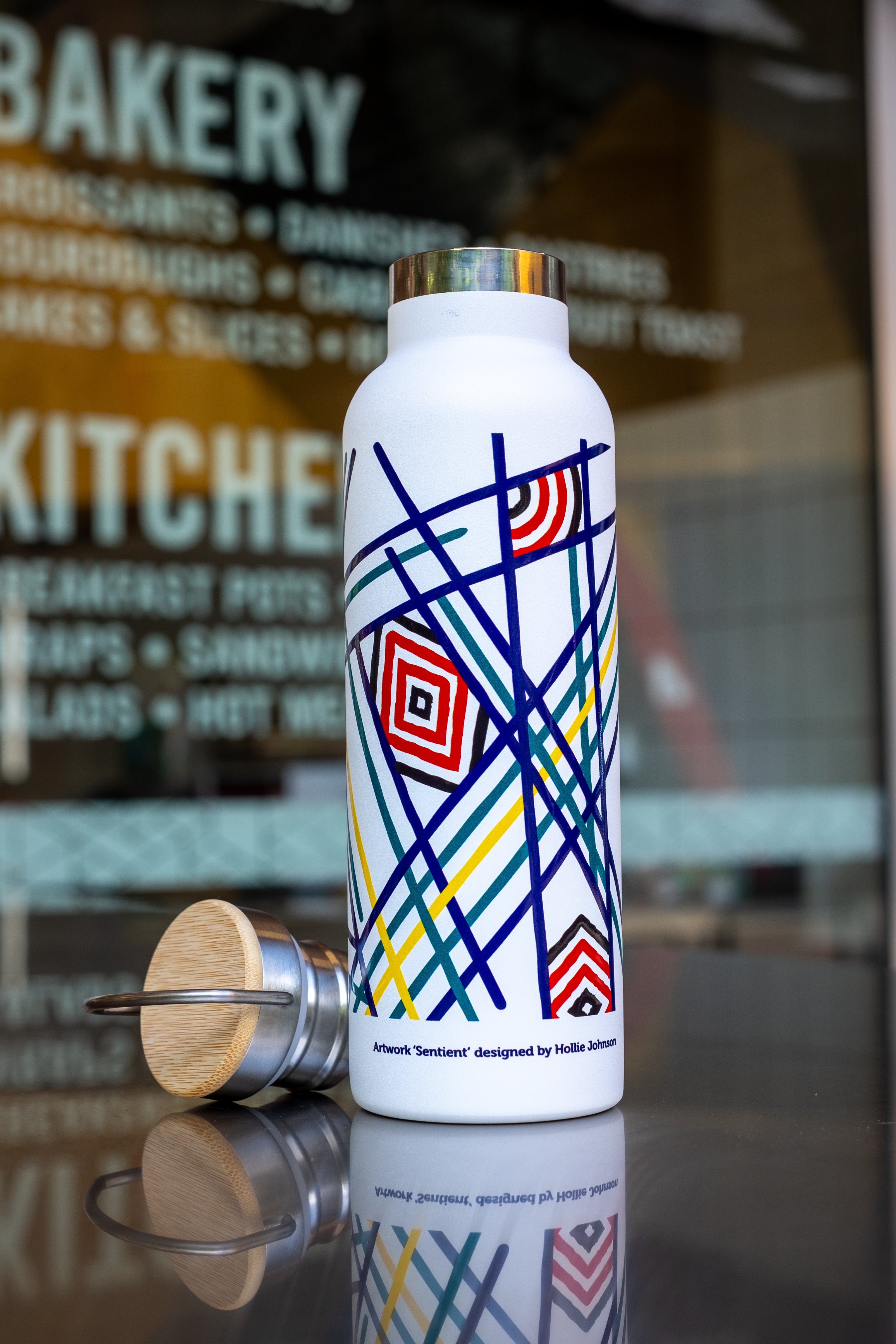White thermo bottle with colourful geometric artwork 'Sentient' by Hollie Johnson, featuring a detachable wooden lid, displayed on a reflective surface.