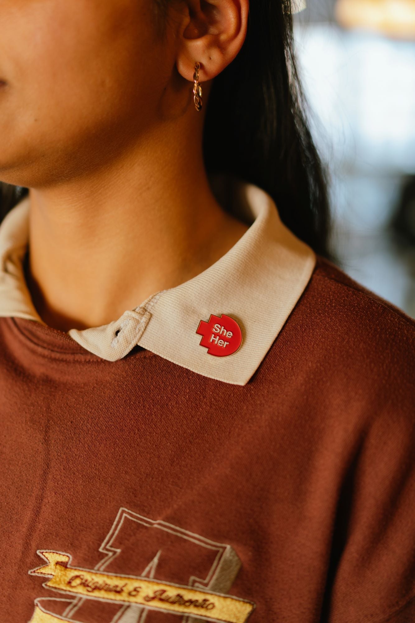 Close-up of a model wearing a red pixelated “she/her” pronoun pin from the RMIT Store, attached to the white collar of her shirt. The pin features white printed text on a red pixelated background. 