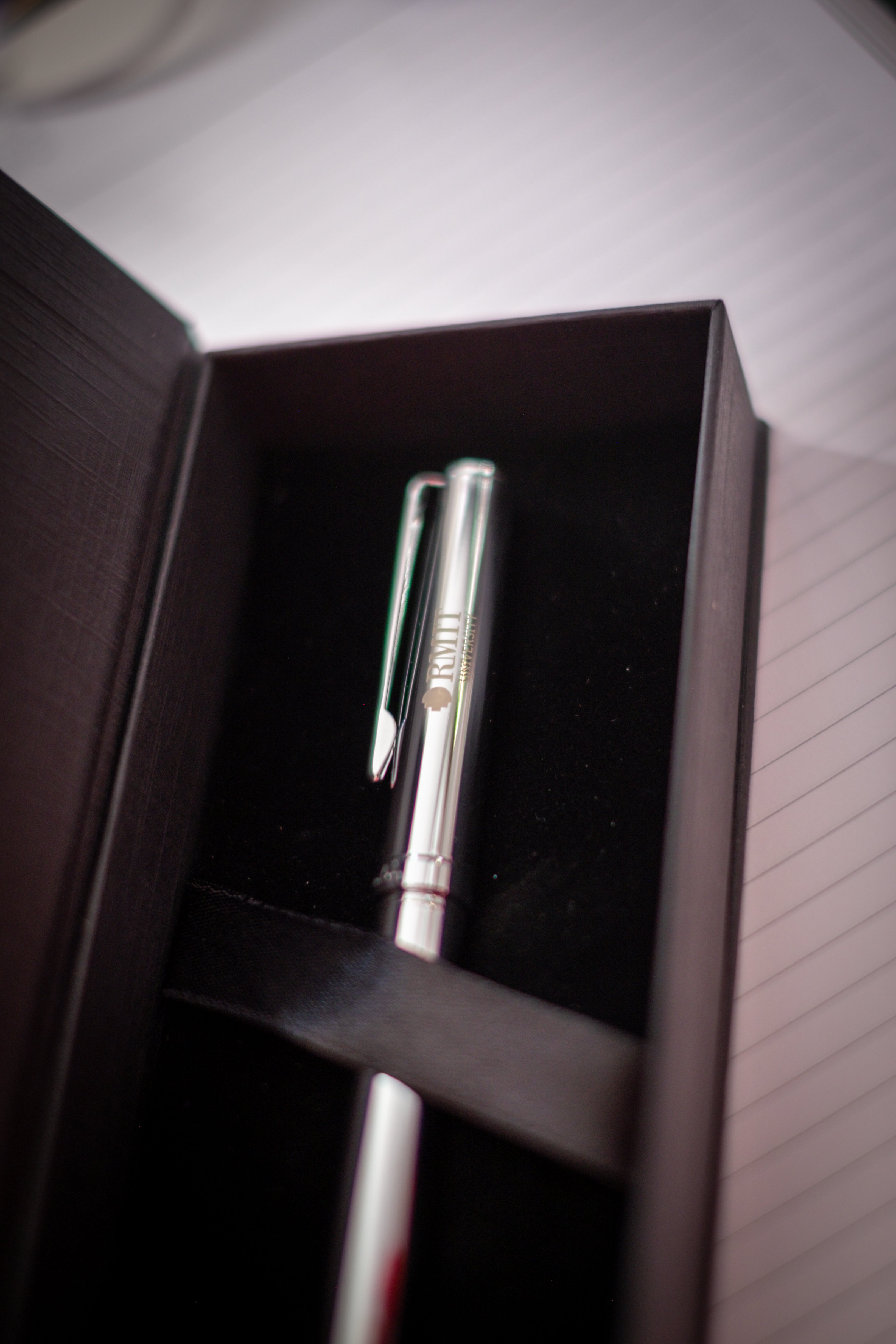 Chrome RMIT Signature Pen with engraved RMIT University logo presented in a black gift box with ribbon strap, shown on a lined notebook background under soft lighting, available at the RMIT Store.