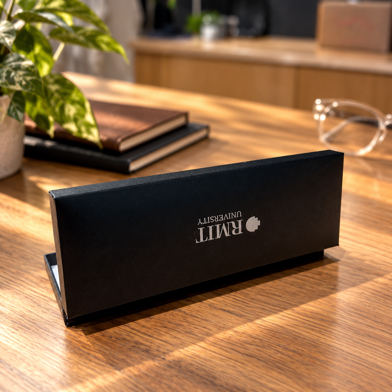 Black textured RMIT Signature Pen gift box with a silver RMIT University logo on the lid, displayed slightly open on a warm wooden desk, with stacked notebooks, a leafy plant and glasses softly blurred in the background. RMIT Store product photography.