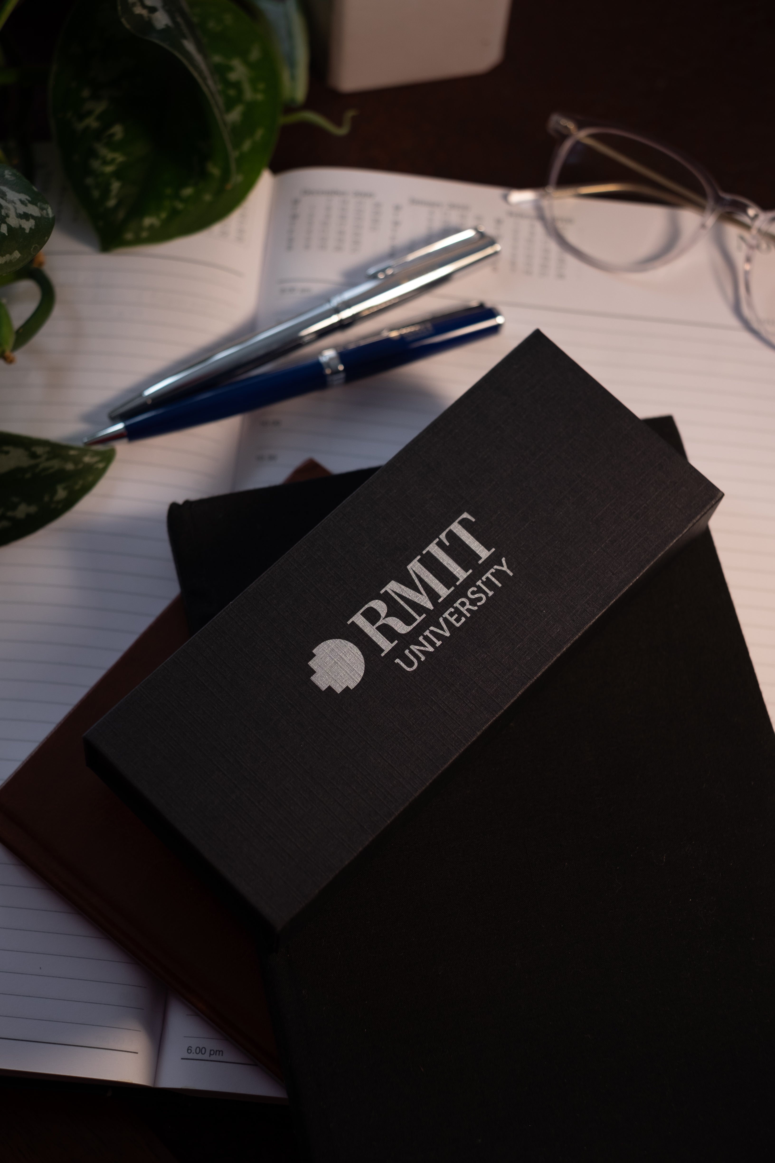 Black RMIT University gift box with silver embossed logo resting on an open planner, with chrome and navy RMIT Signature Pens slightly blurred in the background on a desk setup, available at the RMIT Store.