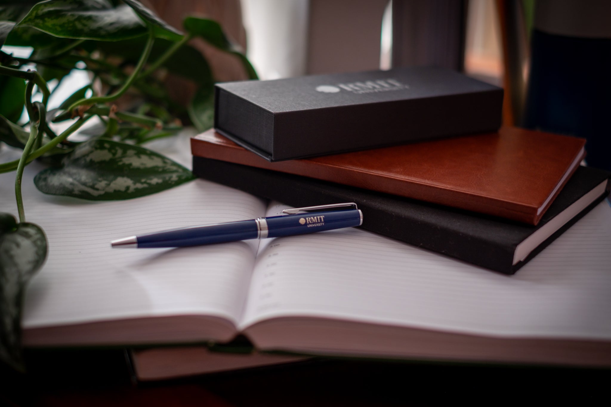 Navy RMIT Signature Pen with silver accents and engraved RMIT University logo resting on an open lined notebook beside stacked brown and black journals and a black RMIT gift box, with green plant leaves framing the scene, available at the RMIT Store.