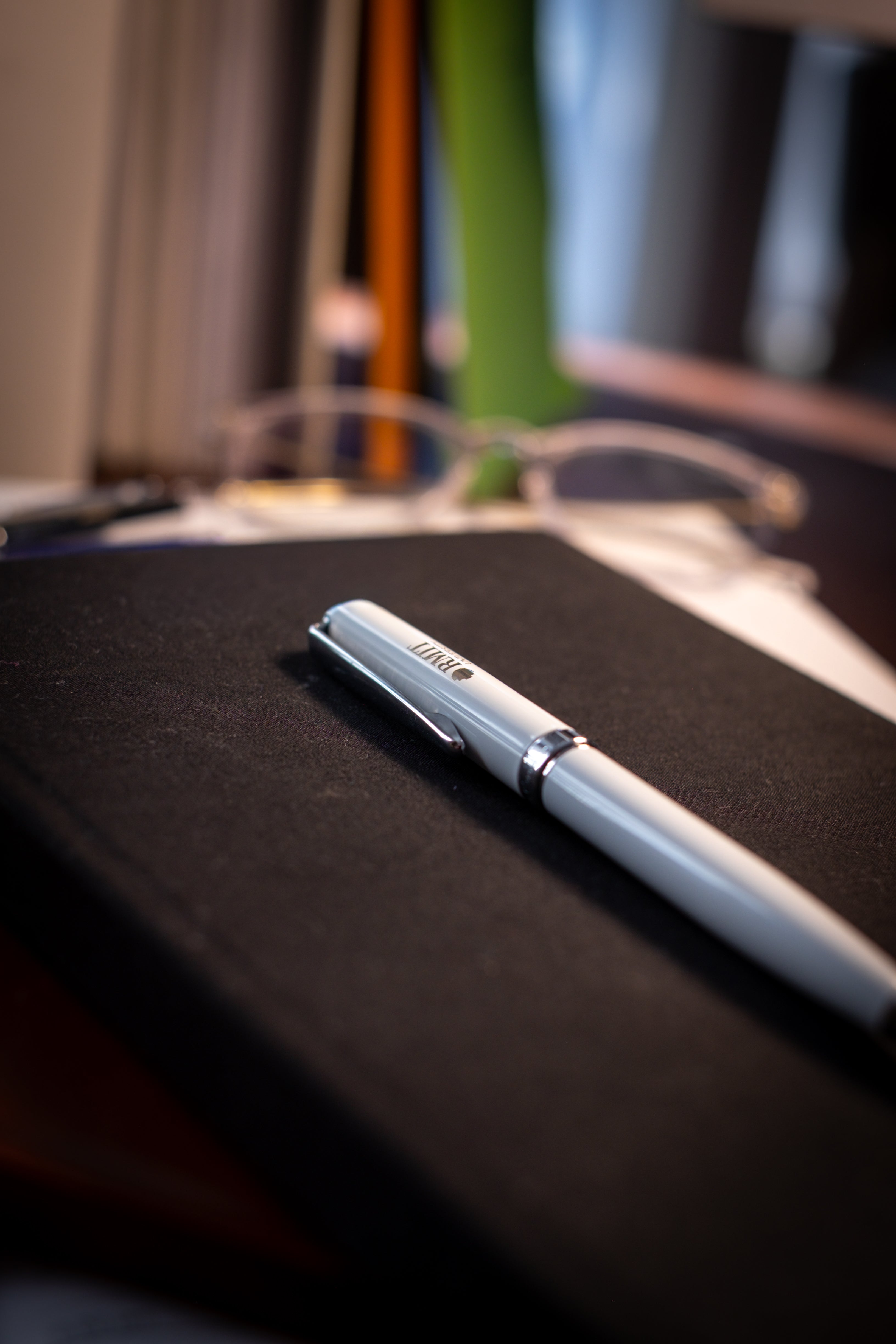 White RMIT Signature Pen with silver detailing and engraved RMIT University logo resting on a black notebook, with glasses and desk accessories blurred in the background, available at the RMIT Store.