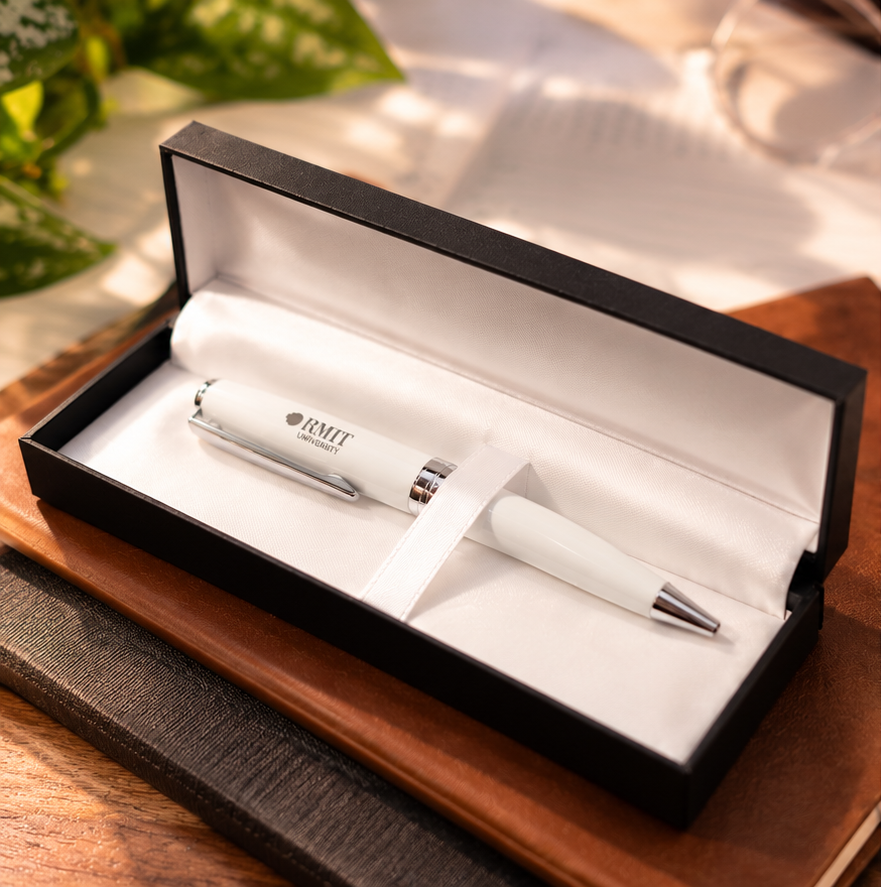 White RMIT Signature Pen with silver-coloured clip, centre band and tip, featuring a small grey RMIT University logo on the barrel. Presented inside an open black gift box with white satin-style lining and a white ribbon holding the pen in place, resting on stacked brown notebooks in warm natural light. RMIT Store product photography.