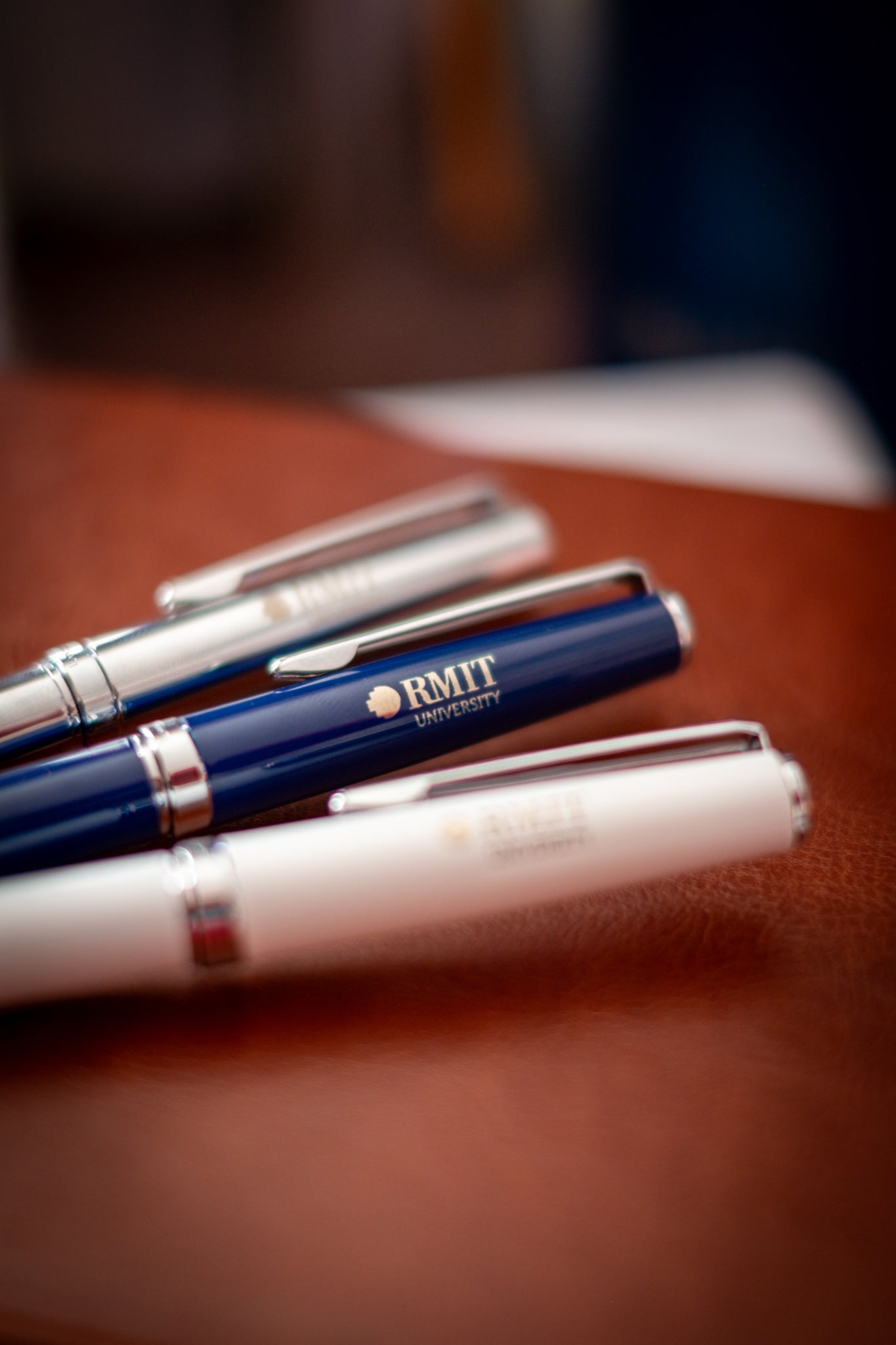 Three RMIT Signature Pens in chrome, navy, and white variants arranged diagonally on a light brown leather surface, each featuring the RMIT University logo engraved near the clip, photographed in soft studio lighting for the RMIT Store.