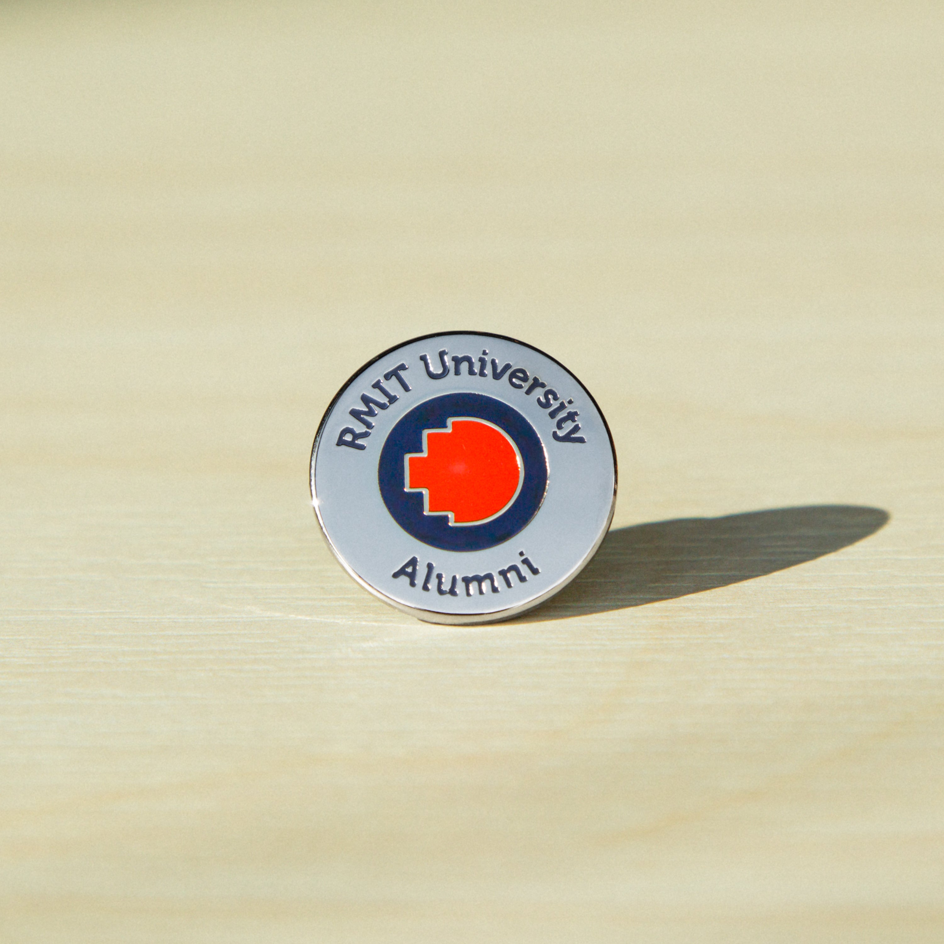 Close-up of a silver metal RMIT alumni pin standing upright on a wooden tabletop. The pin features an engraved “RMIT University Alumni” text around the edge of the round pin, with the RMIT logo prominently centred. The polished metal surface reflects soft natural light.