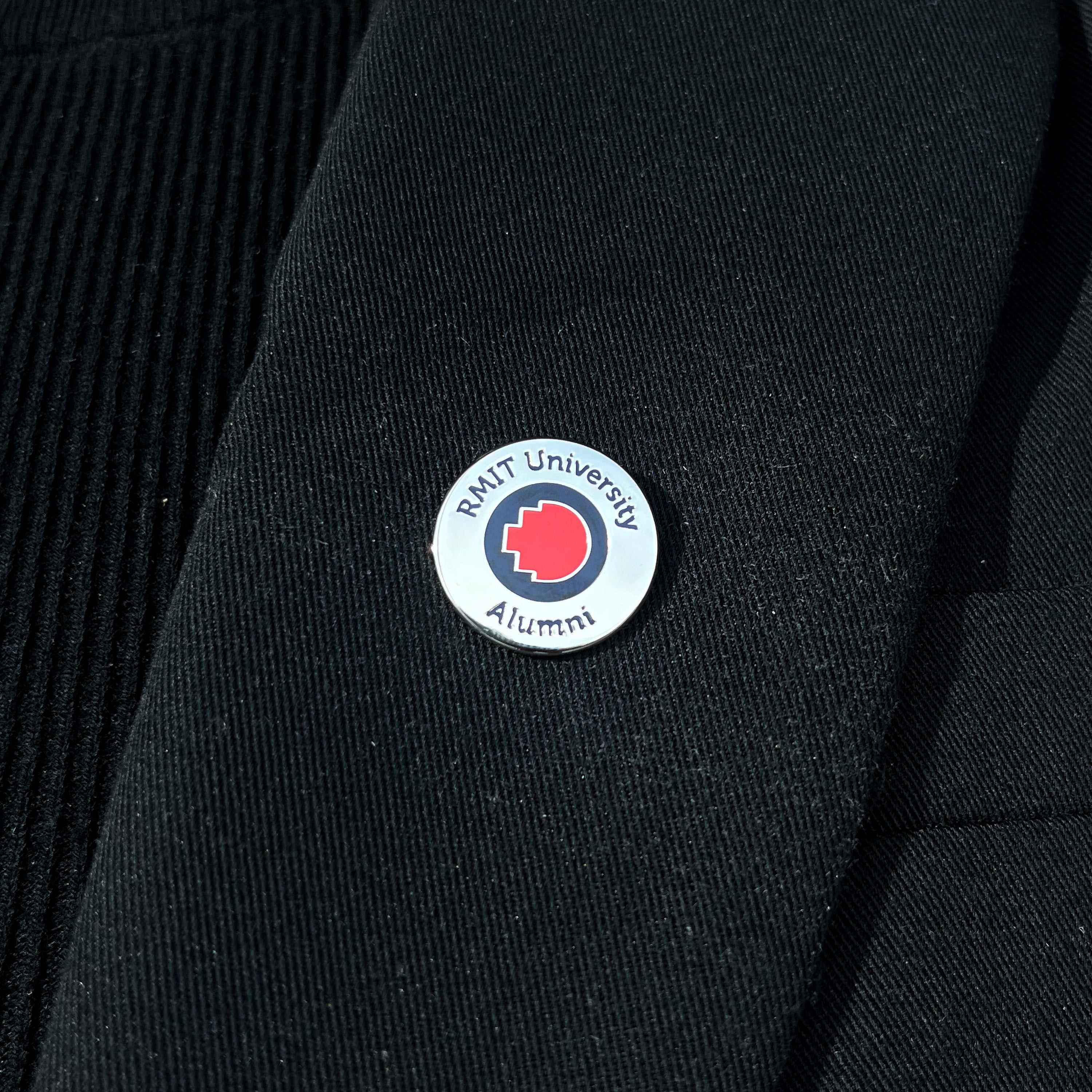 Close-up of a silver metal RMIT alumni pin attached to the lapel of a black blazer. The pin features a subtle embossed RMIT logo and a polished finish, shown against the fabric texture of the blazer.