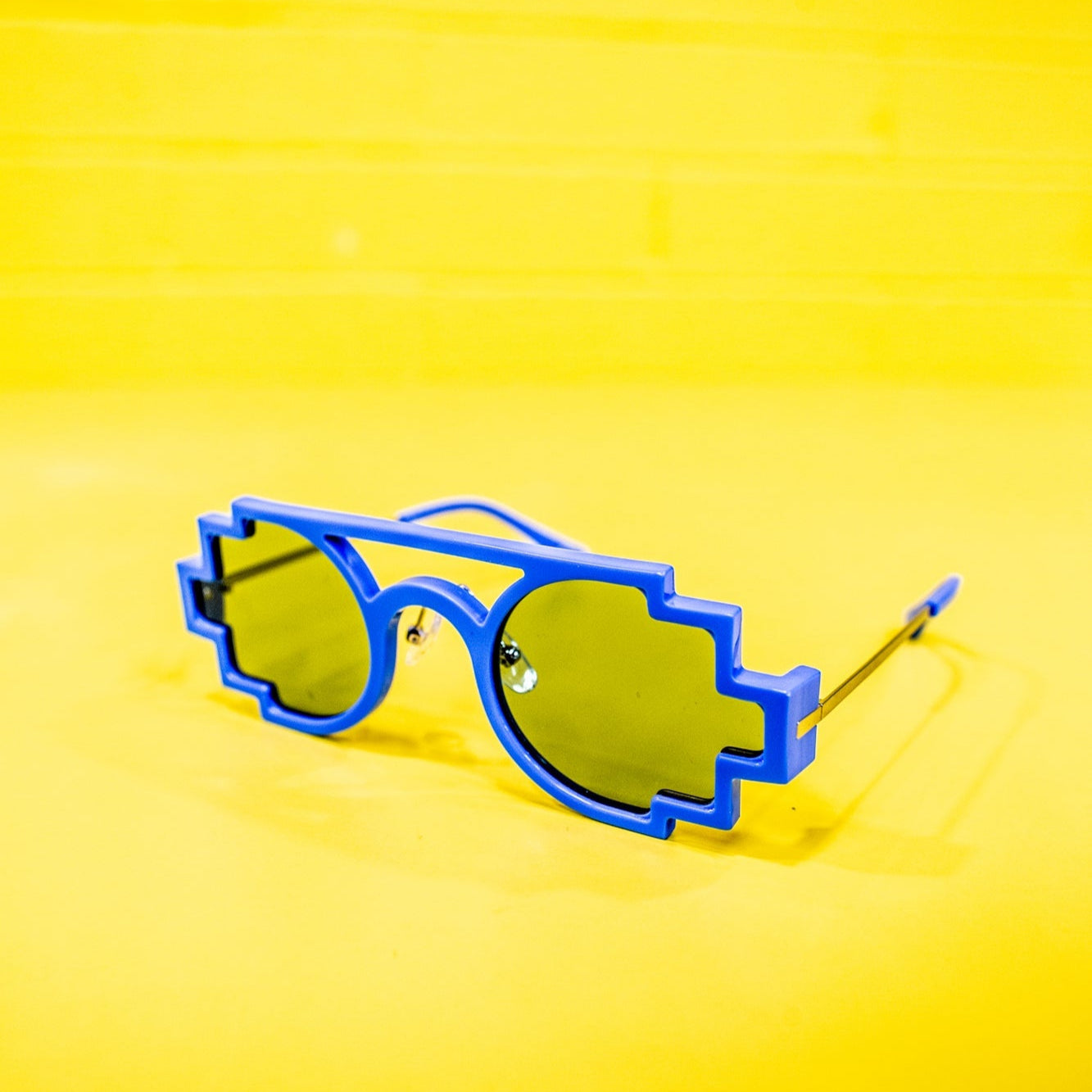 Full front view of sky blue pixelated sunglasses featuring a blocky, pixel-inspired design with squared lenses and thick frames. The sunglasses have a matte finish and are positioned against a yellow studio background for the RMIT Store.