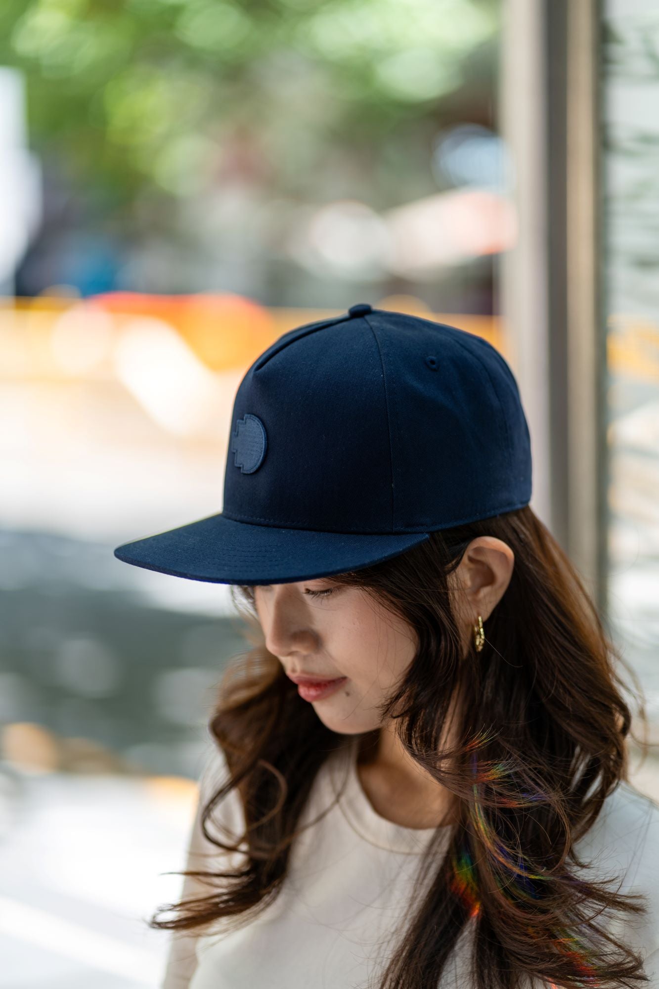 RMIT Store navy blue snapback cap with an embroidered logo, styled by a model outdoors. The urban scene and rainbow light add vibrancy to the casual, modern apparel.