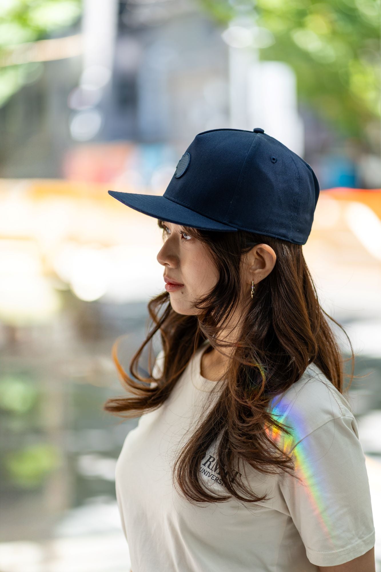 Side view of RMIT Store snapback cap in navy blue with embroidered logo, worn by a model with long, wavy hair and gold hoop earrings. The cap complements the casual outfit in an outdoor urban setting.