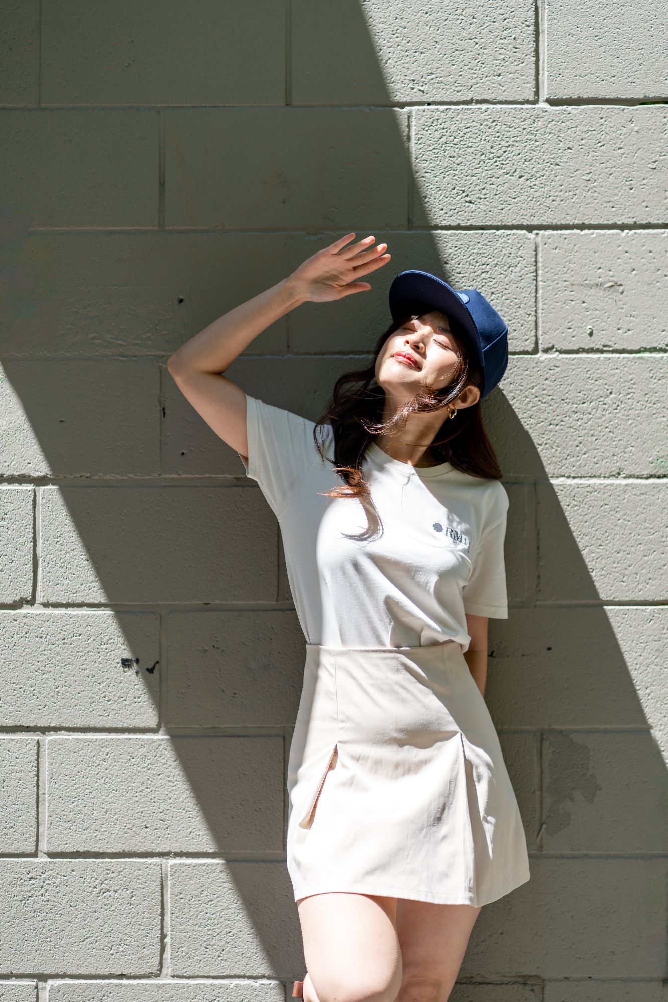 RMIT Store snapback cap in navy blue, paired with a white RMIT University t-shirt and cream skirt. The model poses with one arm raised against a textured wall, showcasing an urban casual style.
