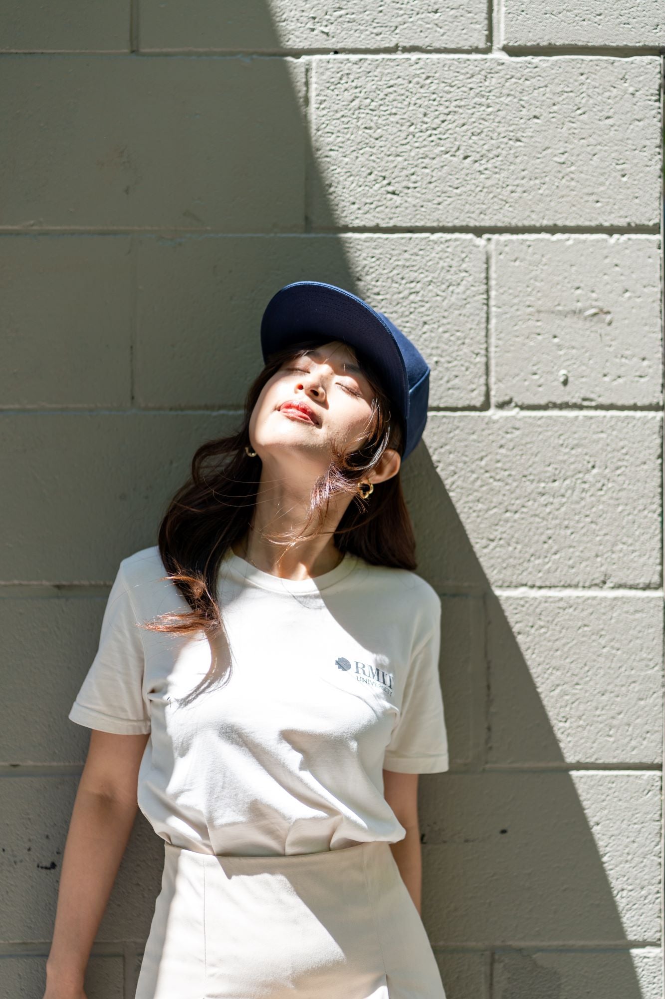 RMIT Store snapback cap in navy blue worn by a model with a white RMIT University t-shirt and cream skirt, standing against a shadowed brick wall. The cap is designed for a stylish, casual look.