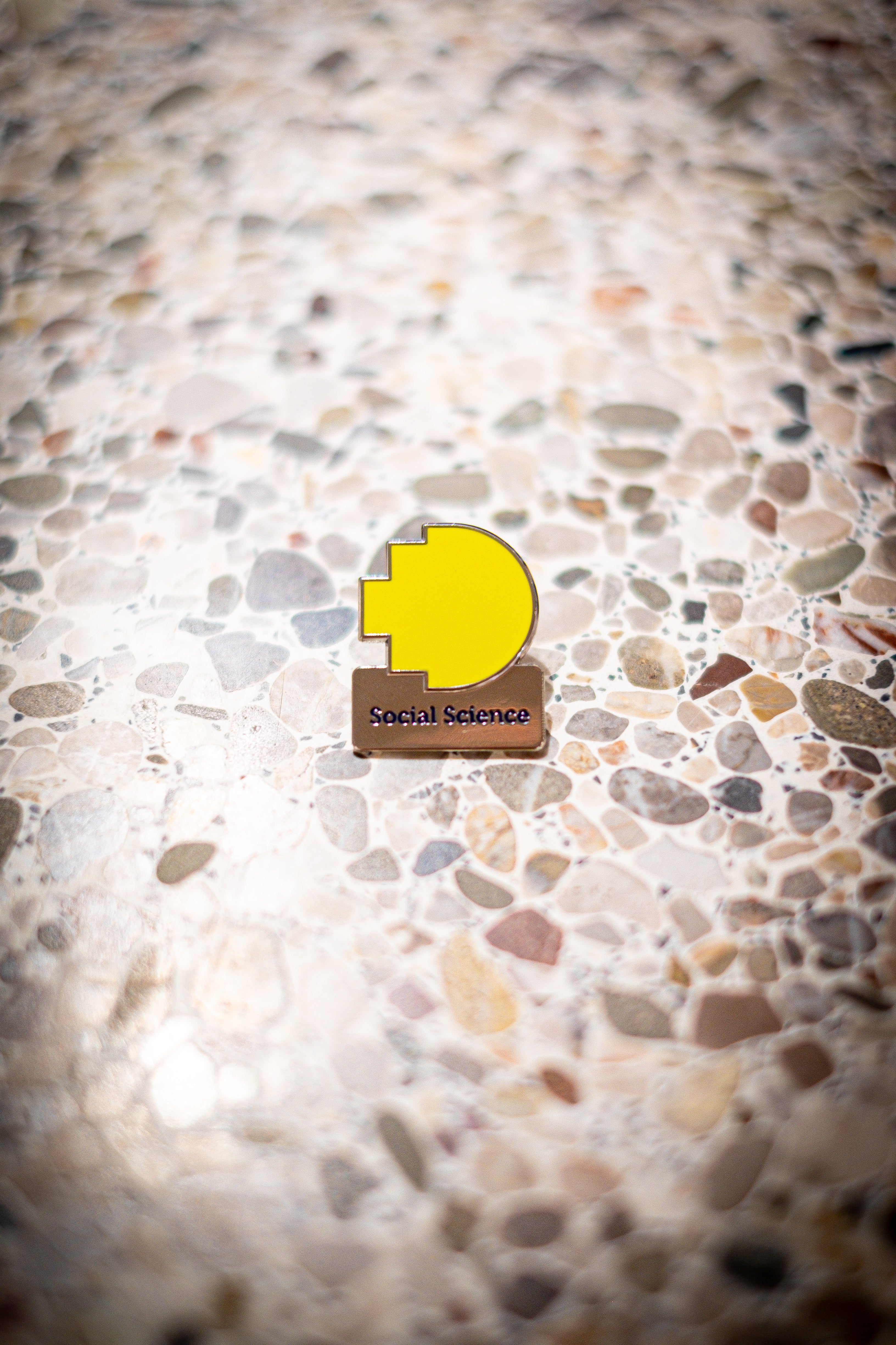 Front view of the Social Science Discipline pin placed on a light marble background, featuring the RMIT pixel logo in yellow above a silver square with the word "Social Science". Available at the RMIT Store.