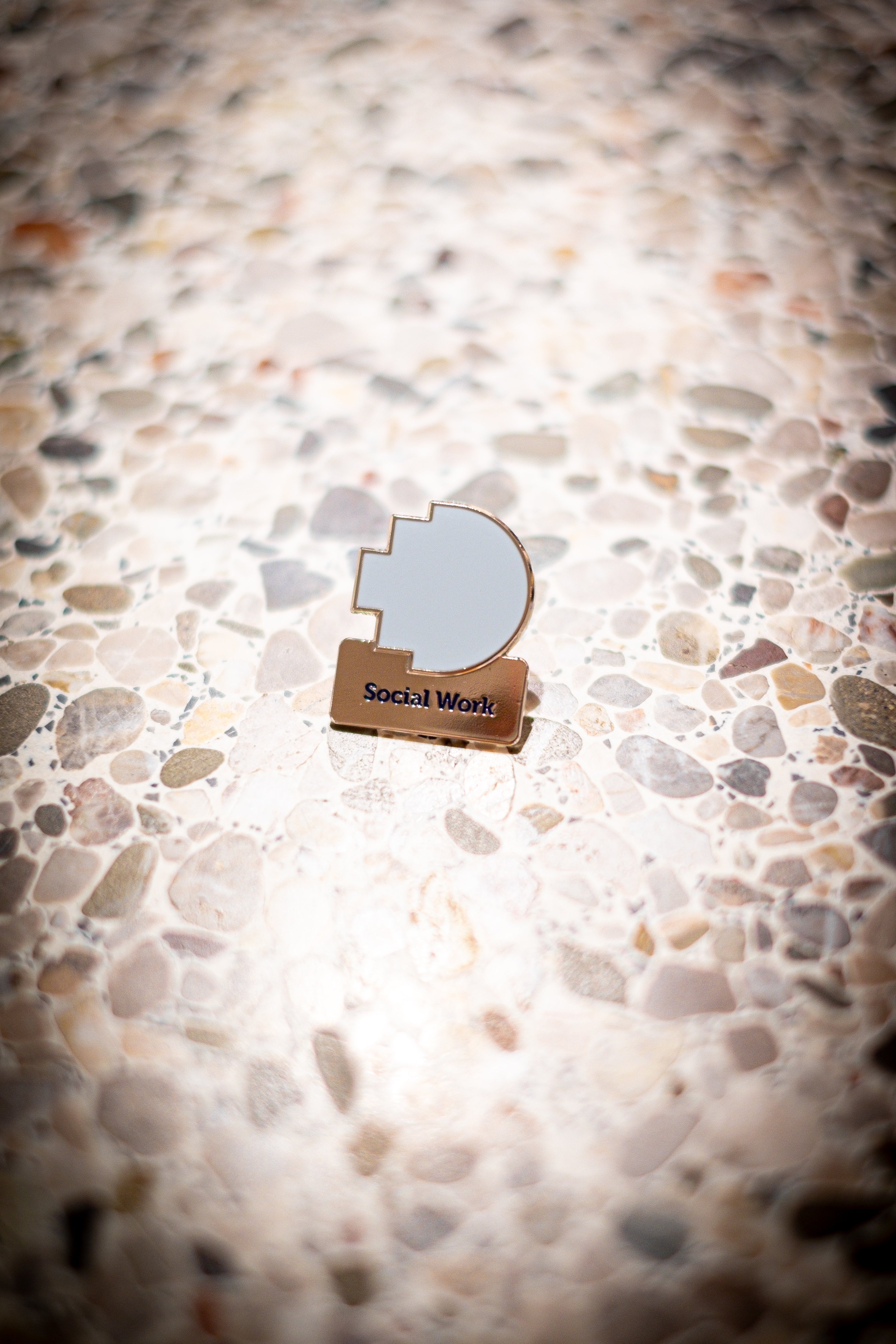 Front view of the Social Work Discipline pin placed on a light marble background, featuring the RMIT pixel logo in light blue above a silver square with the word "Social Work". Available at the RMIT Store.