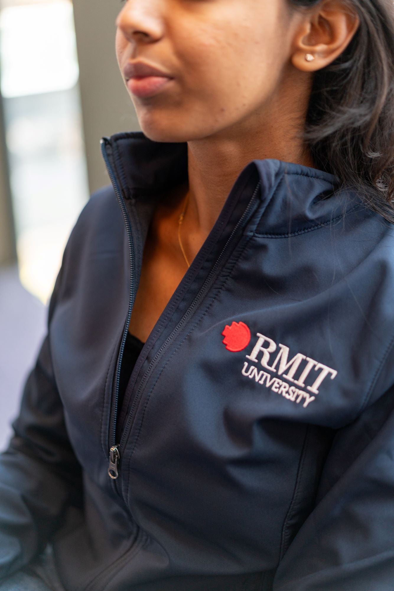 Close-up of the RMIT Softshell Jacket, half unzipped, showcasing the embroidered RMIT logo on the left chest. The model, facing forward, wears the jacket in a studio setting, highlighting its breathable polyester outer layer and high collar.