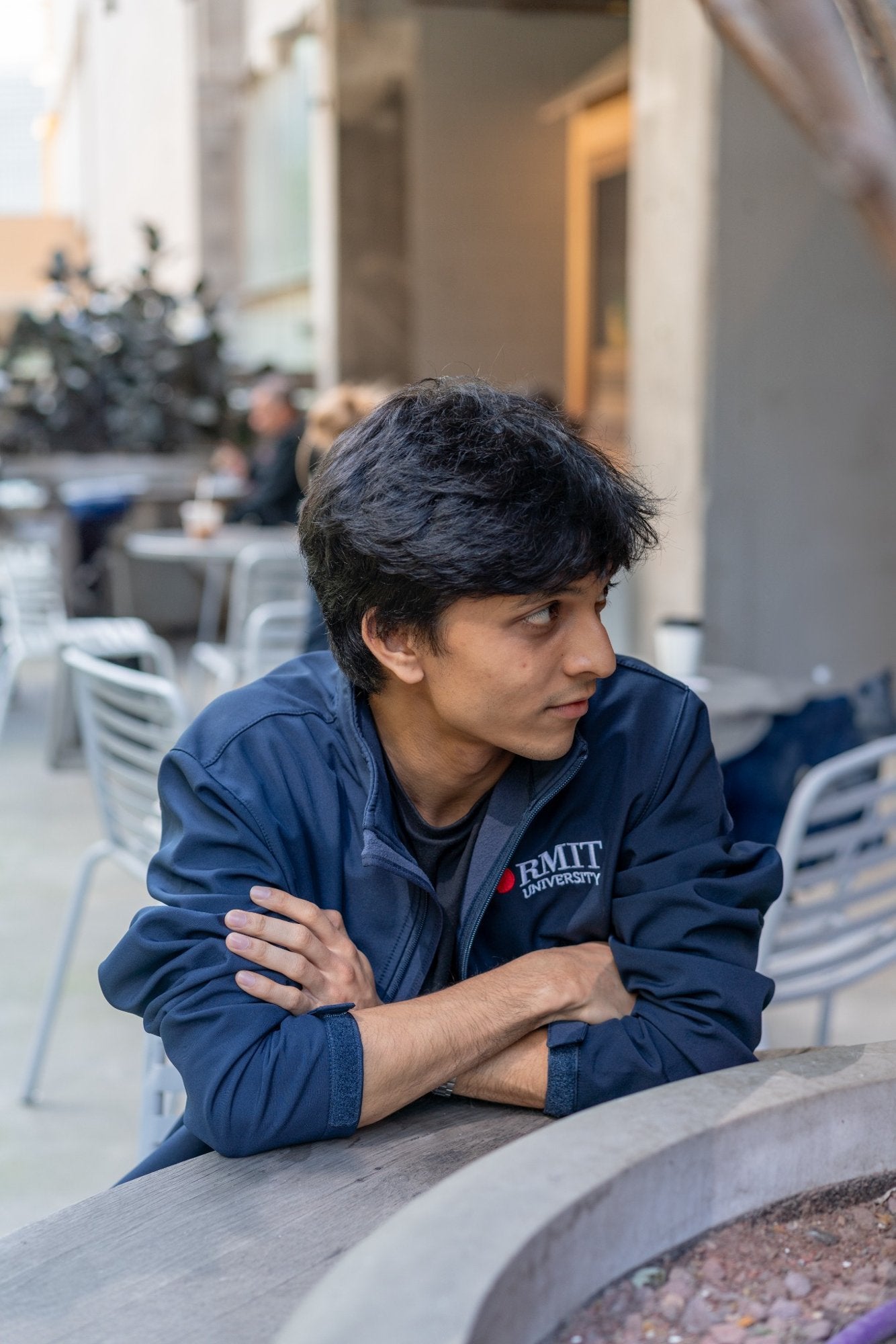 Softshell Jacket in navy worn by a seated male model leaning on an outdoor table, showing the front upper body fit, high collar, long sleeves, cuff tabs, full zip opening, and embroidered RMIT University logo on the left chest with the red RMIT pixel icon and white text, available at the RMIT Store.