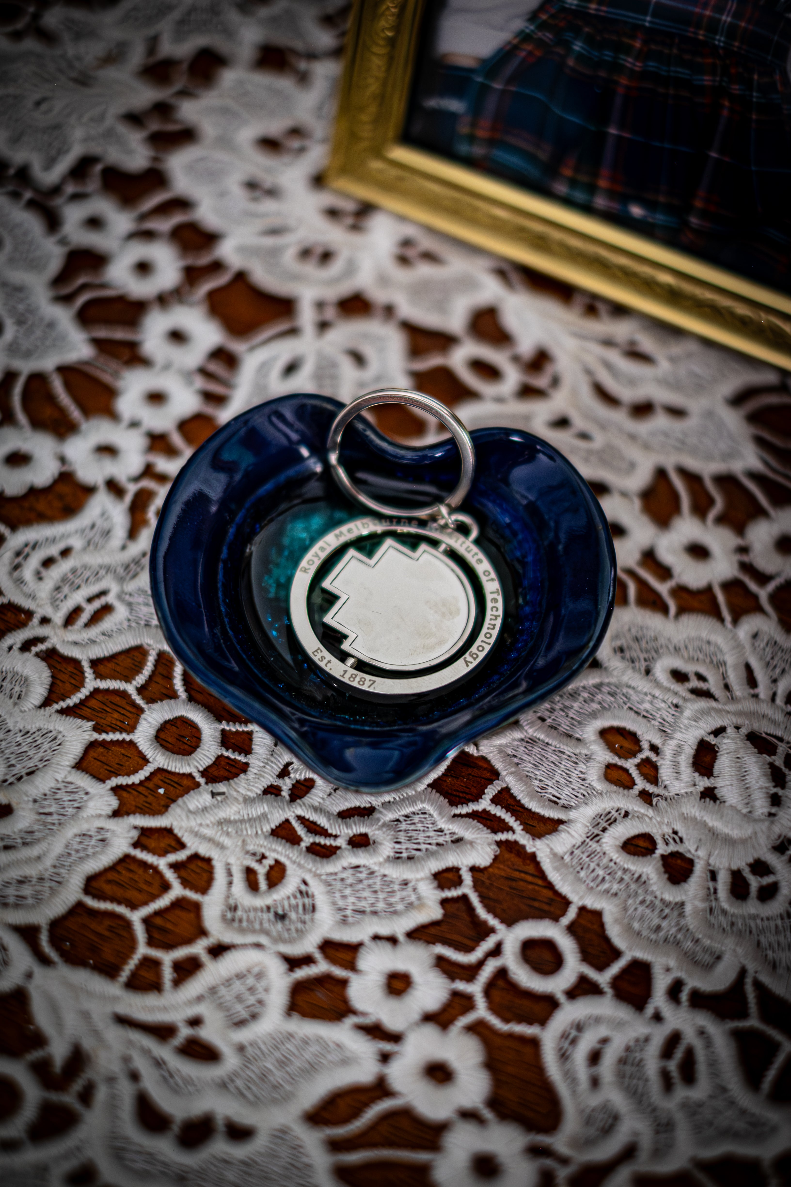 RMIT Spinner Pixel Keyring in polished silver metal, featuring the RMIT pixel shape at the centre and engraved text reading “Royal Melbourne Institute of Technology Est. 1887,” displayed inside a glossy dark blue ceramic dish on a lace-covered wooden surface beside a framed photo. Available at the RMIT Store.