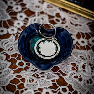 RMIT Spinner Pixel Keyring in polished silver metal, featuring the RMIT pixel shape at the centre and engraved text reading “Royal Melbourne Institute of Technology Est. 1887,” displayed inside a glossy dark blue ceramic dish on a lace-covered wooden surface beside a framed photo. Available at the RMIT Store.
