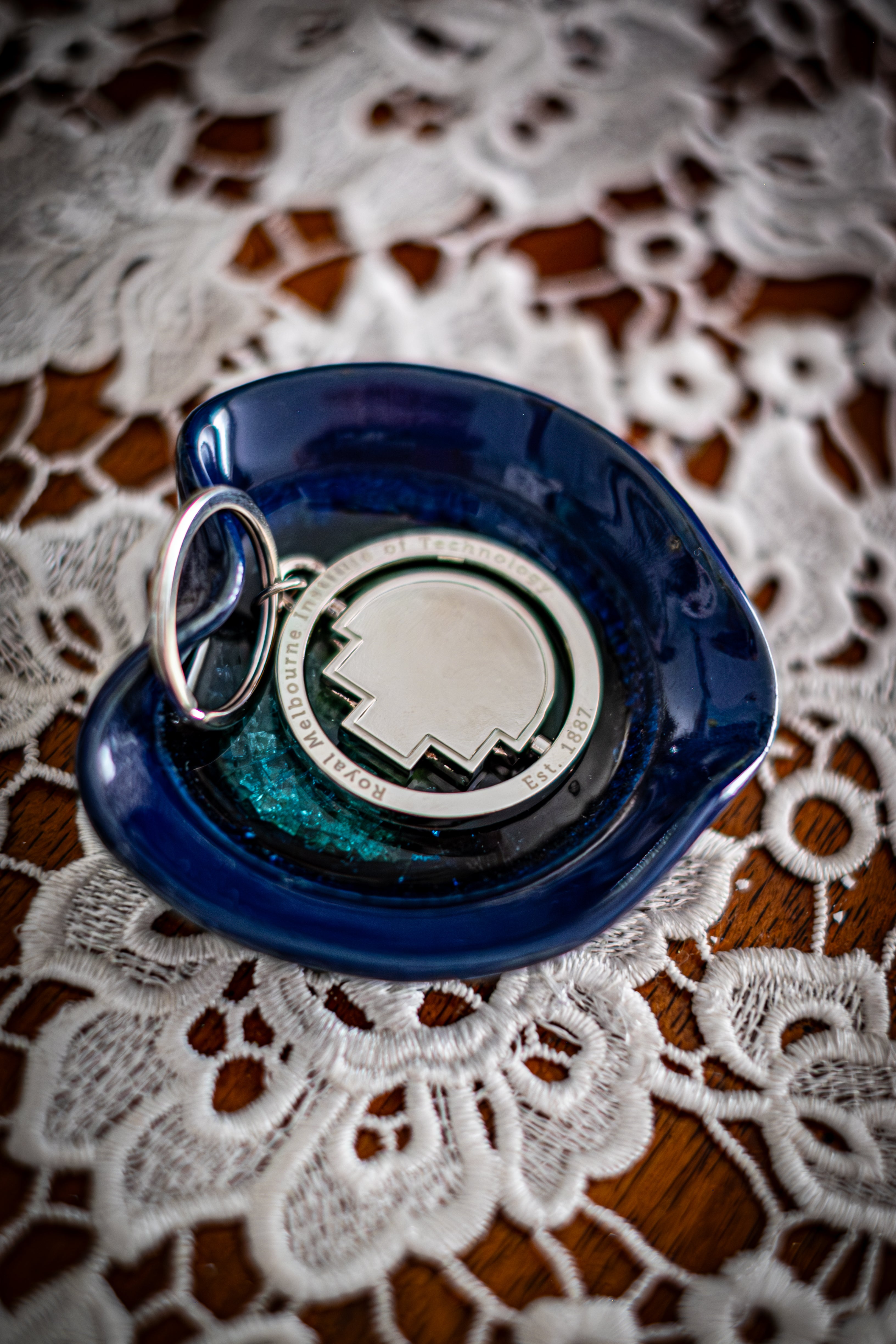 RMIT Spinner Pixel Keyring in polished silver metal, featuring the RMIT pixel shape at the centre and engraved text reading “Royal Melbourne Institute of Technology Est. 1887,” displayed inside a glossy dark blue ceramic dish on a lace-covered wooden surface. Available at the RMIT Store.