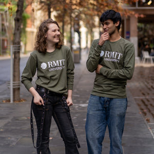 Female and male models wearing Storey Hall L/S Tee tops in Army green featuring bold white RMIT University logo prints on the front, posing together on an outdoor city laneway surrounded by autumn trees and campus-style architecture, available at the RMIT Store.