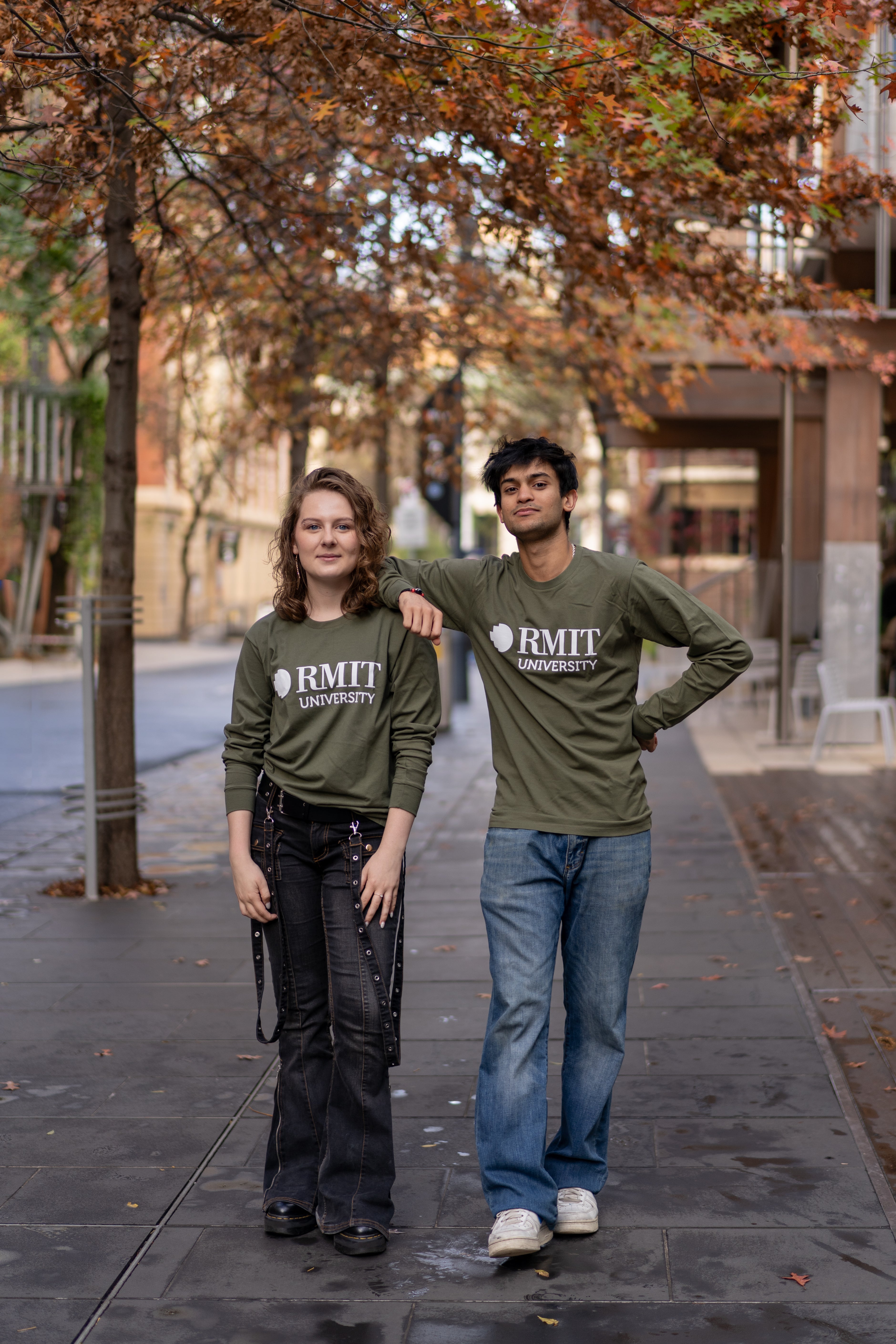Female and male models wearing matching Storey Hall L/S Tee tops in Army green with large white RMIT University chest logos, styled with denim jeans while standing on a tree-lined Melbourne laneway with autumn leaves and urban campus background, available at the RMIT Store.