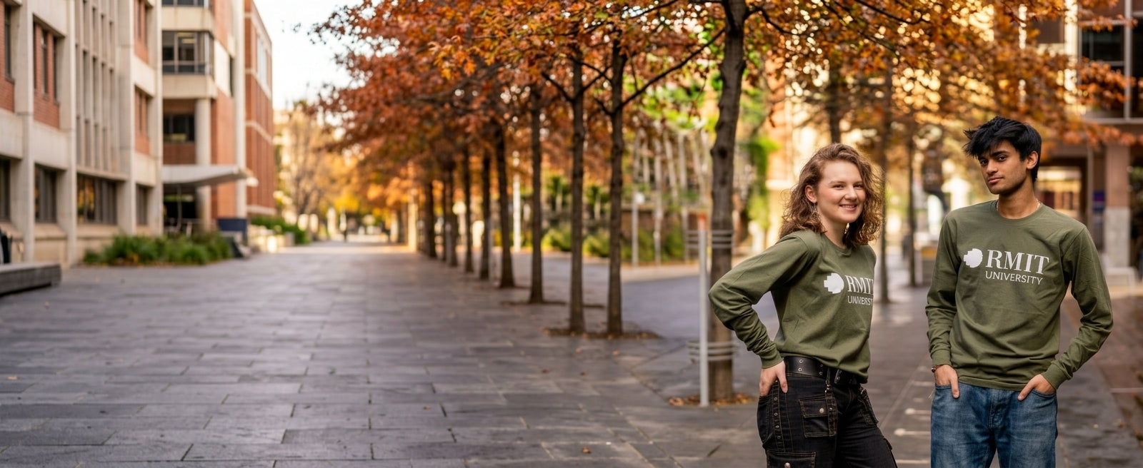Two models wearing Storey Hall long-sleeve tees in an army green colour, each featuring a large white RMIT University logo printed across the chest; one model wears black trousers with belt and strap detailing and the other wears blue jeans, posed on the right of a wide RMIT campus walkway lined with trees and buildings for RMIT Store.
