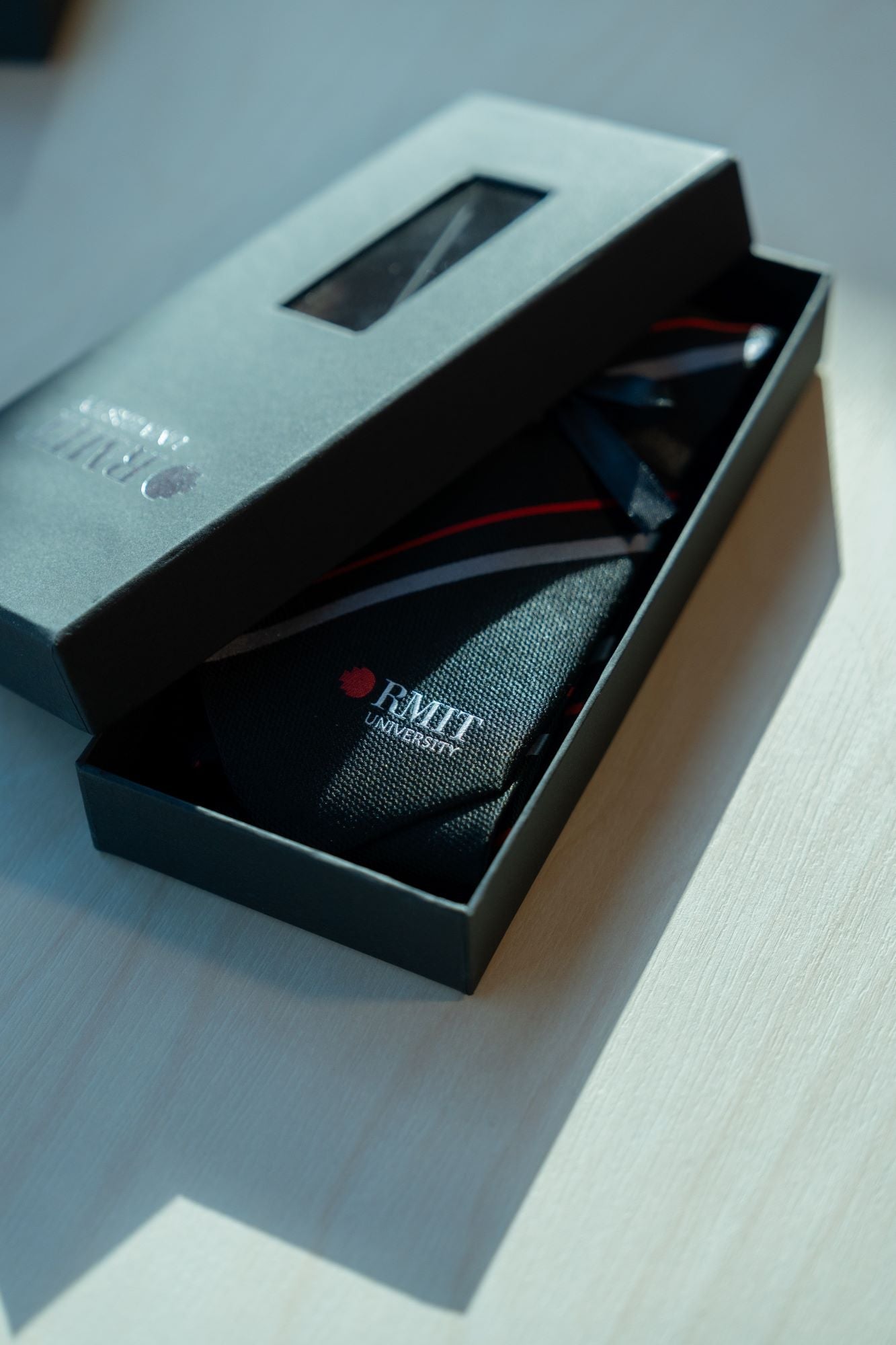 RMIT Stripe Tie presented in a sleek black box with a clear window, highlighting the RMIT University logo in red and white. The tie features diagonal red and grey stripes, adding a professional touch. Available for purchase at the RMIT Store.