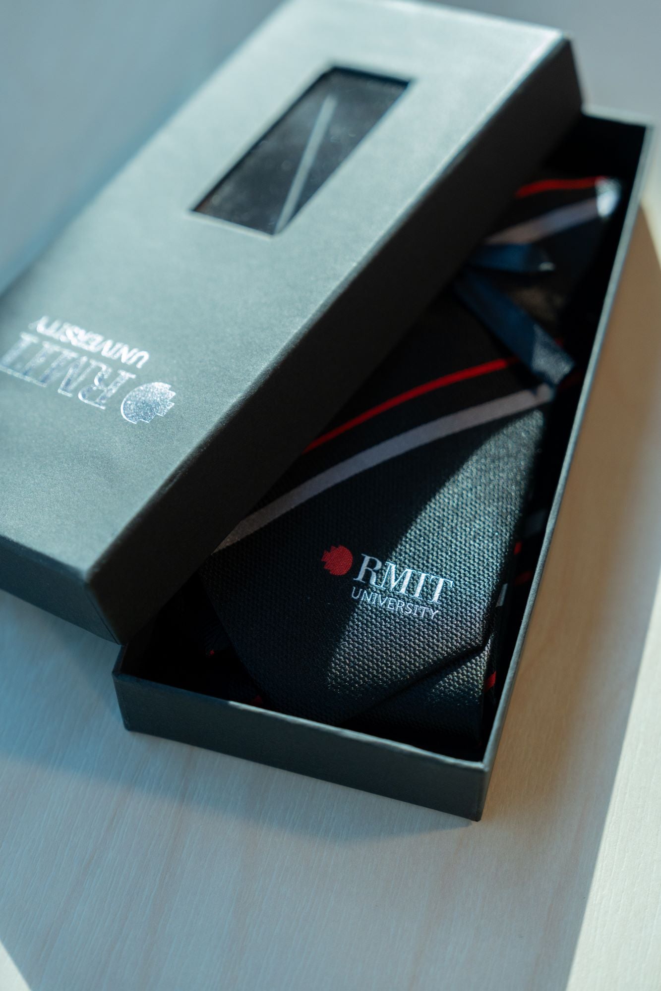 Close-up of the RMIT Stripe Tie inside a presentation box, showing the intricate red and grey stripes and the RMIT University logo. Perfect for formal or academic events, this item is available at the RMIT Store.