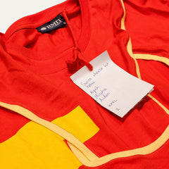 Swiss Cheese But Yellow fashion-student designed upcycled tee from RMIT Store, featuring a red upcycled short-sleeve tee with bright yellow piping tracing cut-out and curved seam lines, yellow rectangular appliqués and dark contrast panels near the hem, shown in a close-up showing the attached white swing tag and nearby reconstructed fabric details on a light studio background.