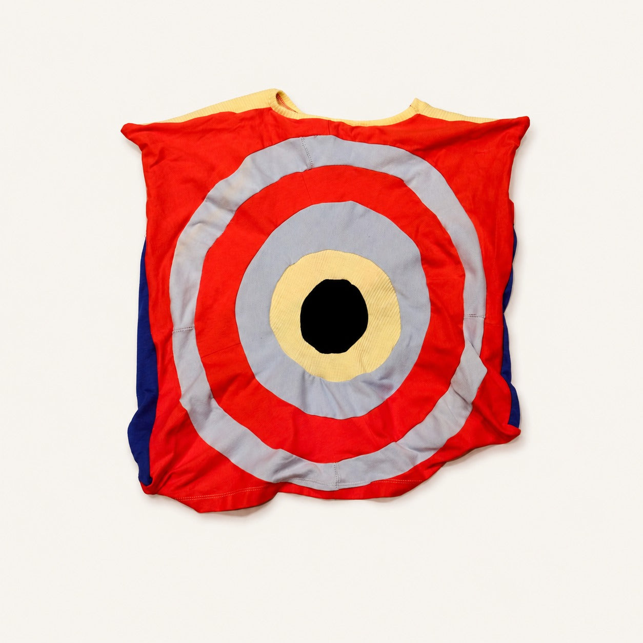 Target On My Back fashion-student designed upcycled tee from RMIT Store, featuring a red upcycled tee with a bold concentric target motif on the back in red, pale blue, cream, black and yellow, with a reconstructed front featuring blue, black, cream and red panels, shown in a rear flat-lay view showing the complete reconstructed silhouette on a light studio background.